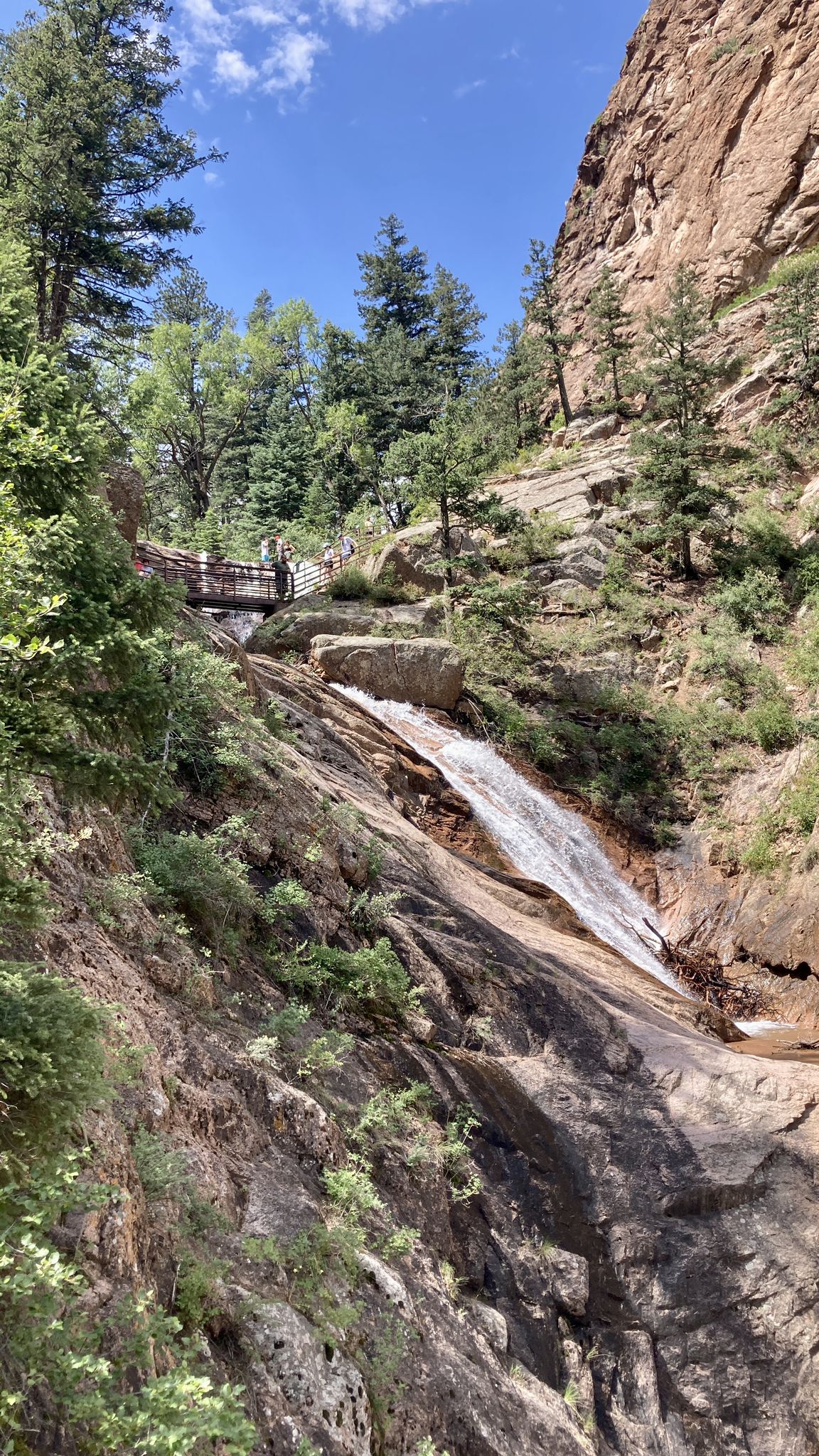 Waterfall cascades down a rocky canyon, with green trees and a bridge above. - Seven Falls to Inspiration Point