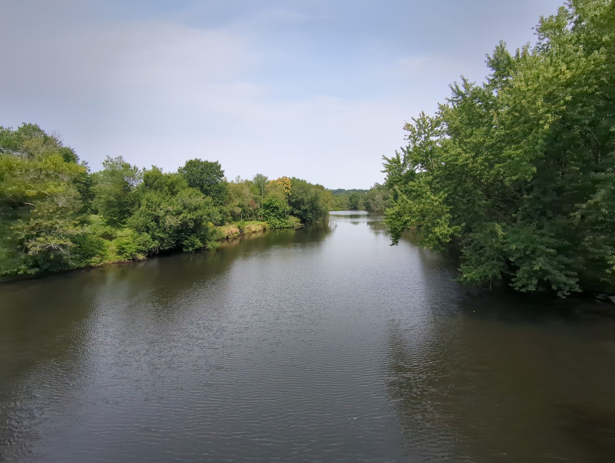 A wide river flows between dense green trees under a bright sky. - Blackstone River Bikeway – Blackstone River Bikeway loop from Providence