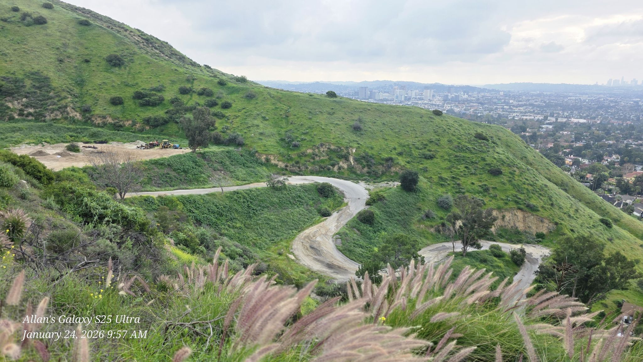 Winding dirt road on a green hillside under cloudy sky, city in distance. - Verdugo Crest and Beaudry Motorway Loop