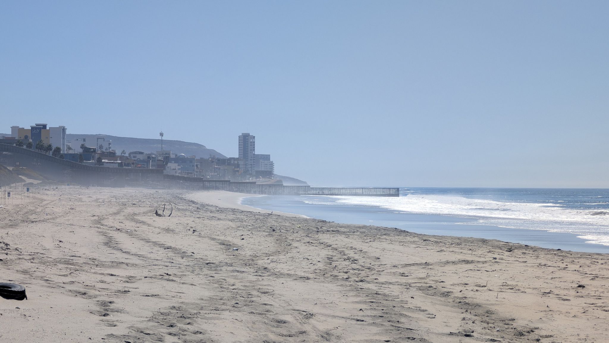 A wide, sandy beach with ocean waves and distant buildings under a hazy sky. - Imperial Beach via Beach Trail & California Coastal Trail