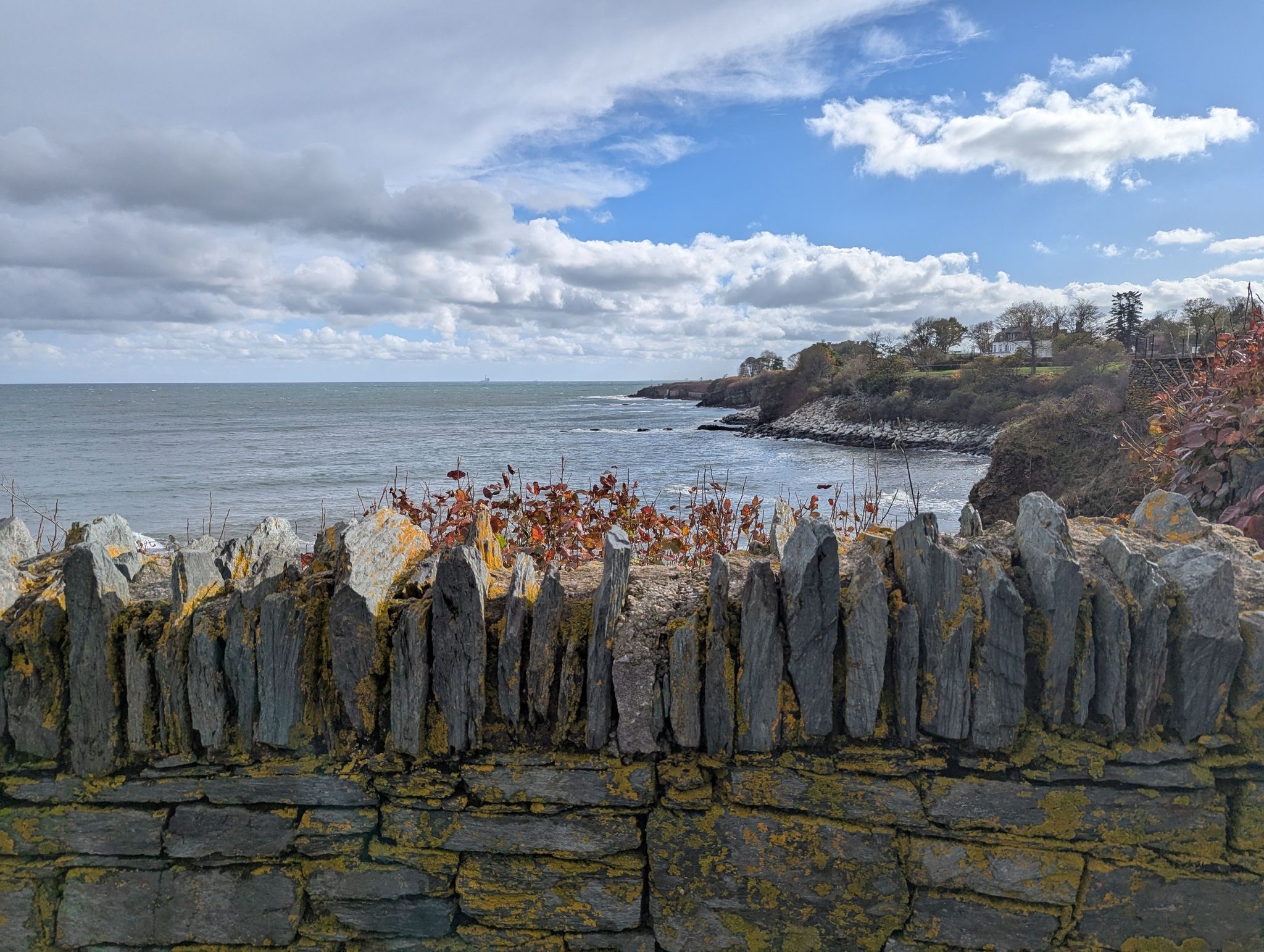 Stone wall overlooks a wavy ocean and distant rocky coastline under clouds. - Forty Steps – View of The Breakers loop from Newport