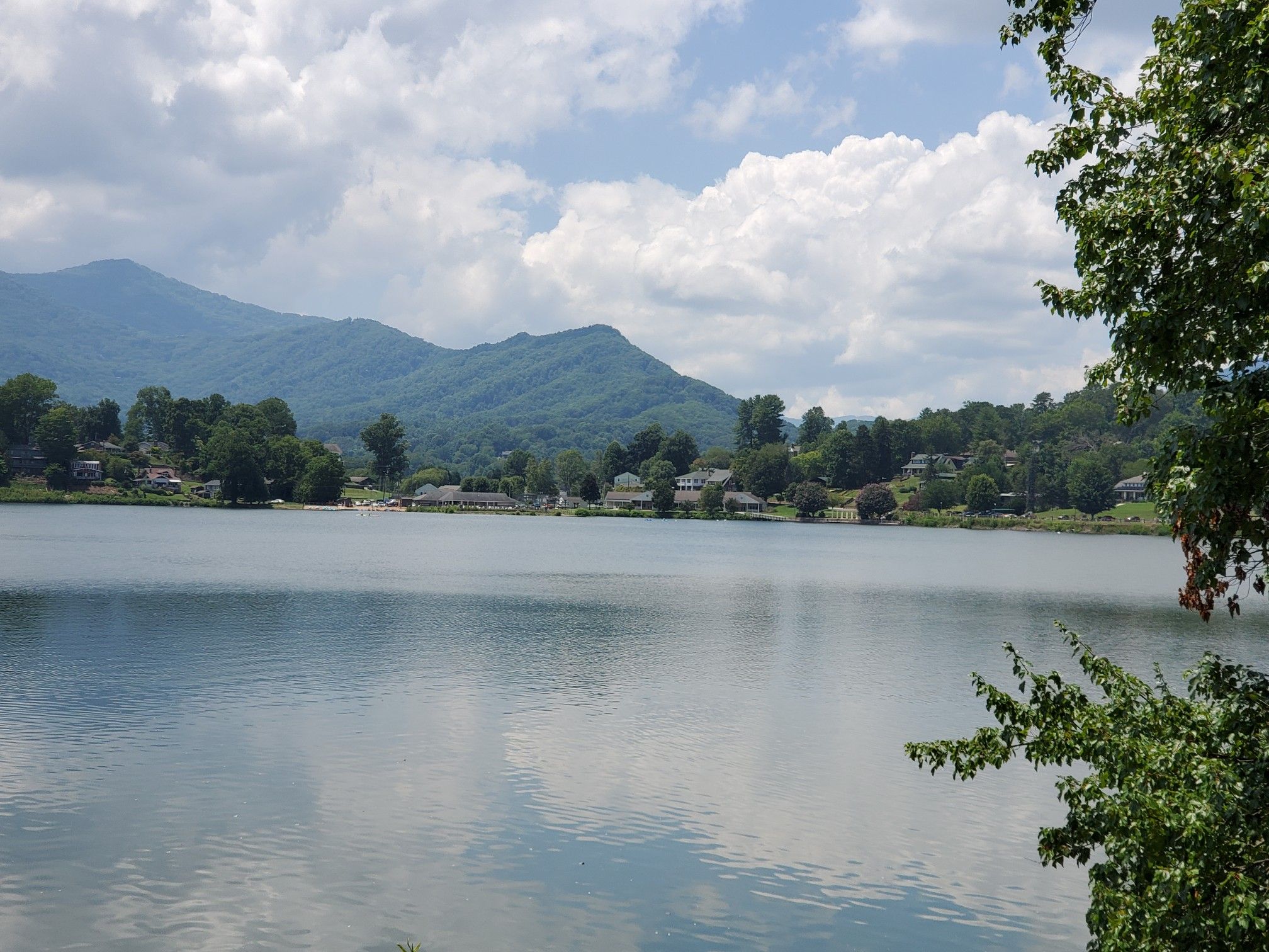 Wide view of a lake with distant mountains and houses under a cloudy sky. - Lake Junaluska Walking Trail