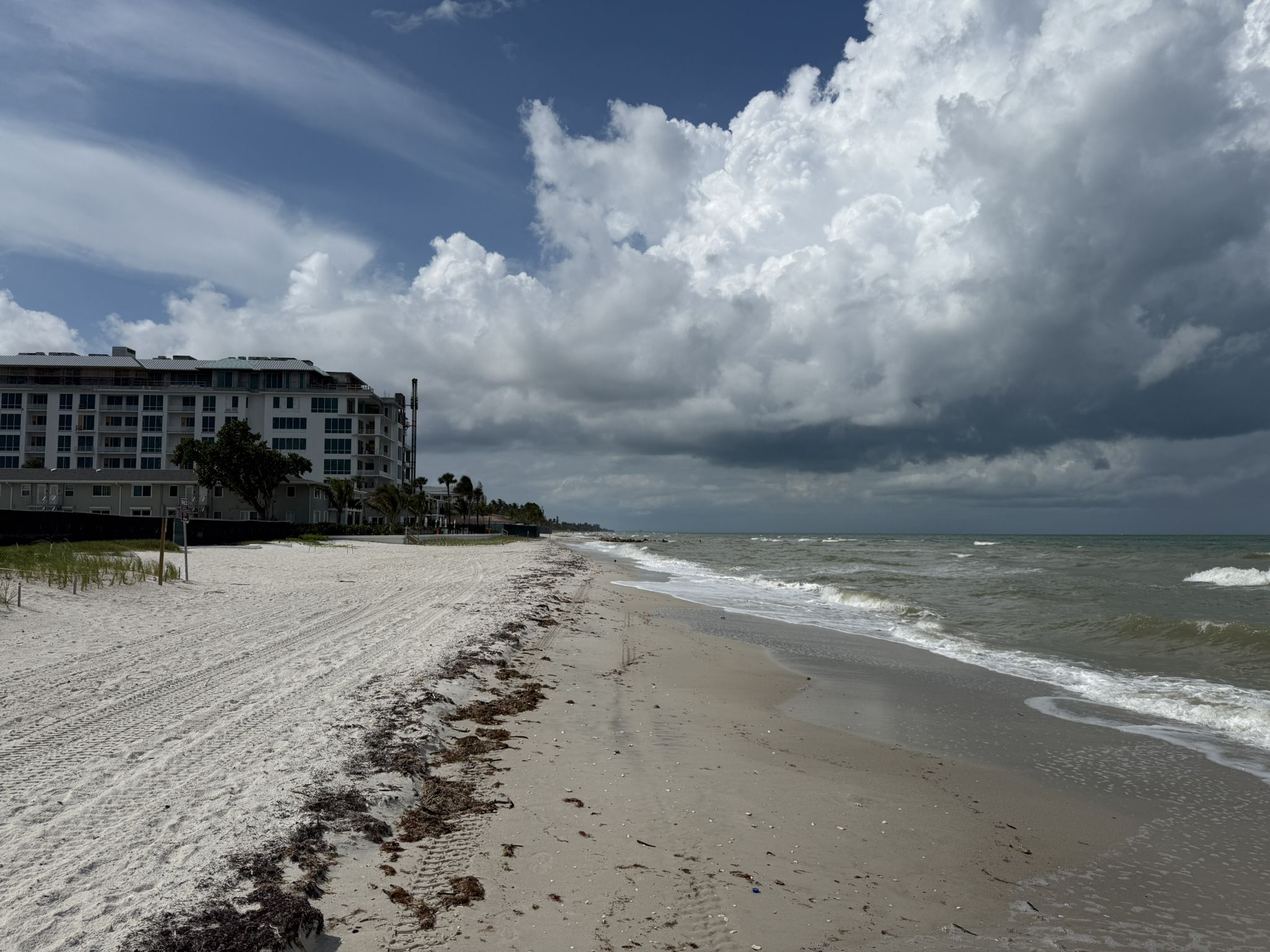 Sandy beach with ocean waves, a large building, and a cloudy sky. - Naples Beach via Lowdermilk Park