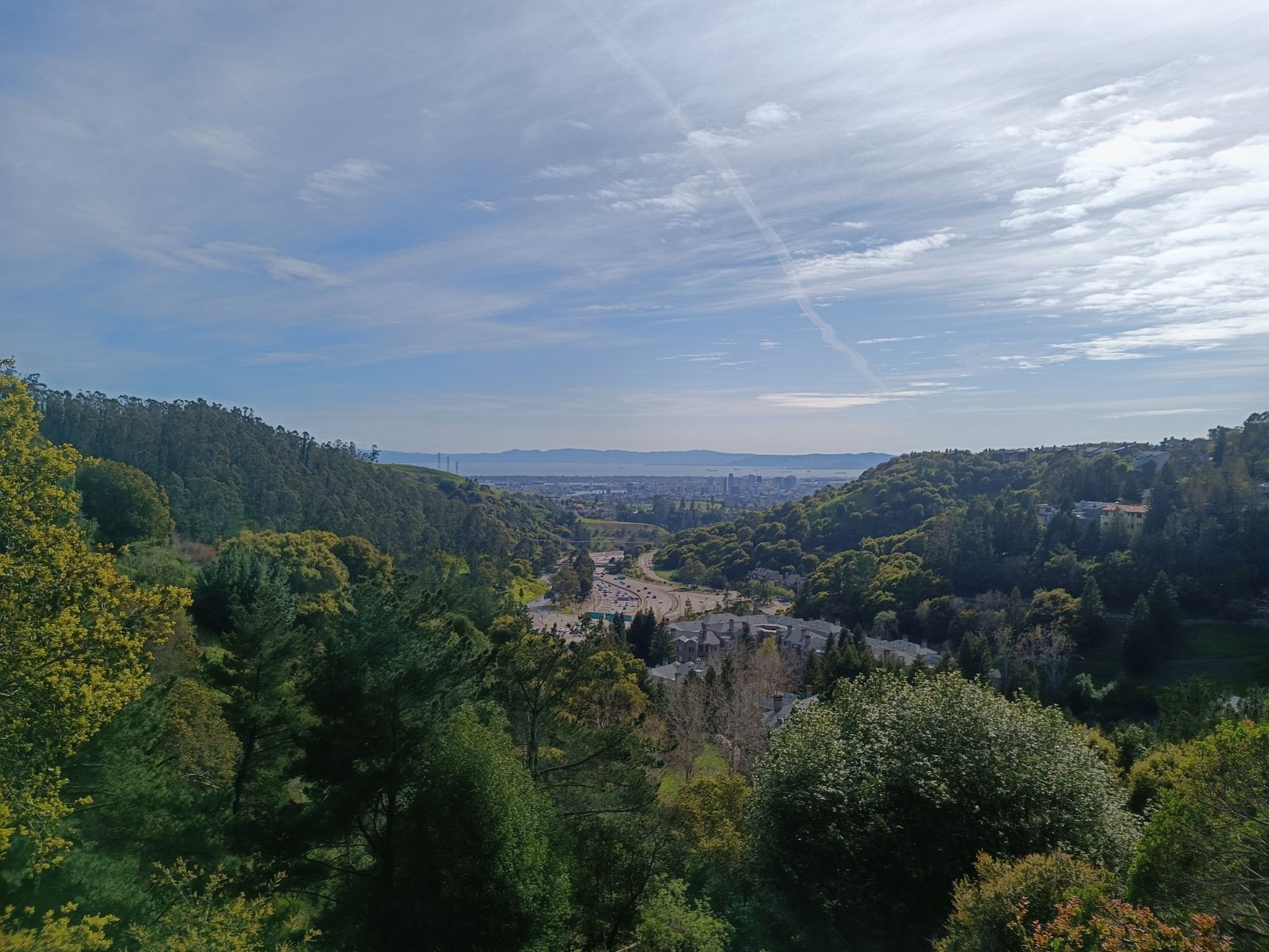 Expansive view of a valley with a city and distant water under a partly cloudy sky. - Clark Kerr Campus, UC Berkeley – Skyline Boulevard loop from North Berkeley