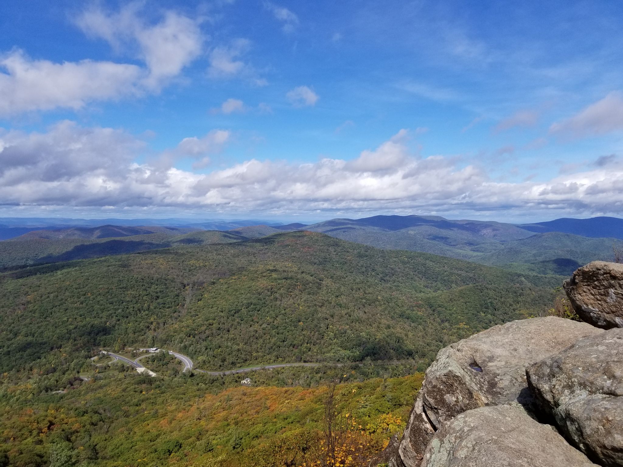 Expansive mountain vista with green forests, a winding road, and cloudy blue sky. - Marys Rock Summit Trail