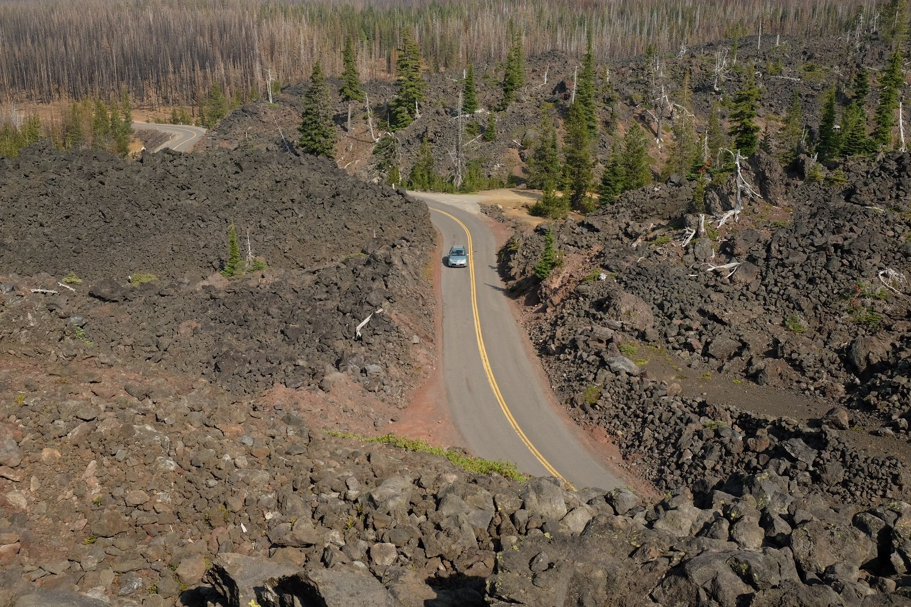 A car drives on a winding road through dark, rocky terrain under bright sun. - Dee Wright Observatory and Lava Trail run — Mount Washington Wilderness