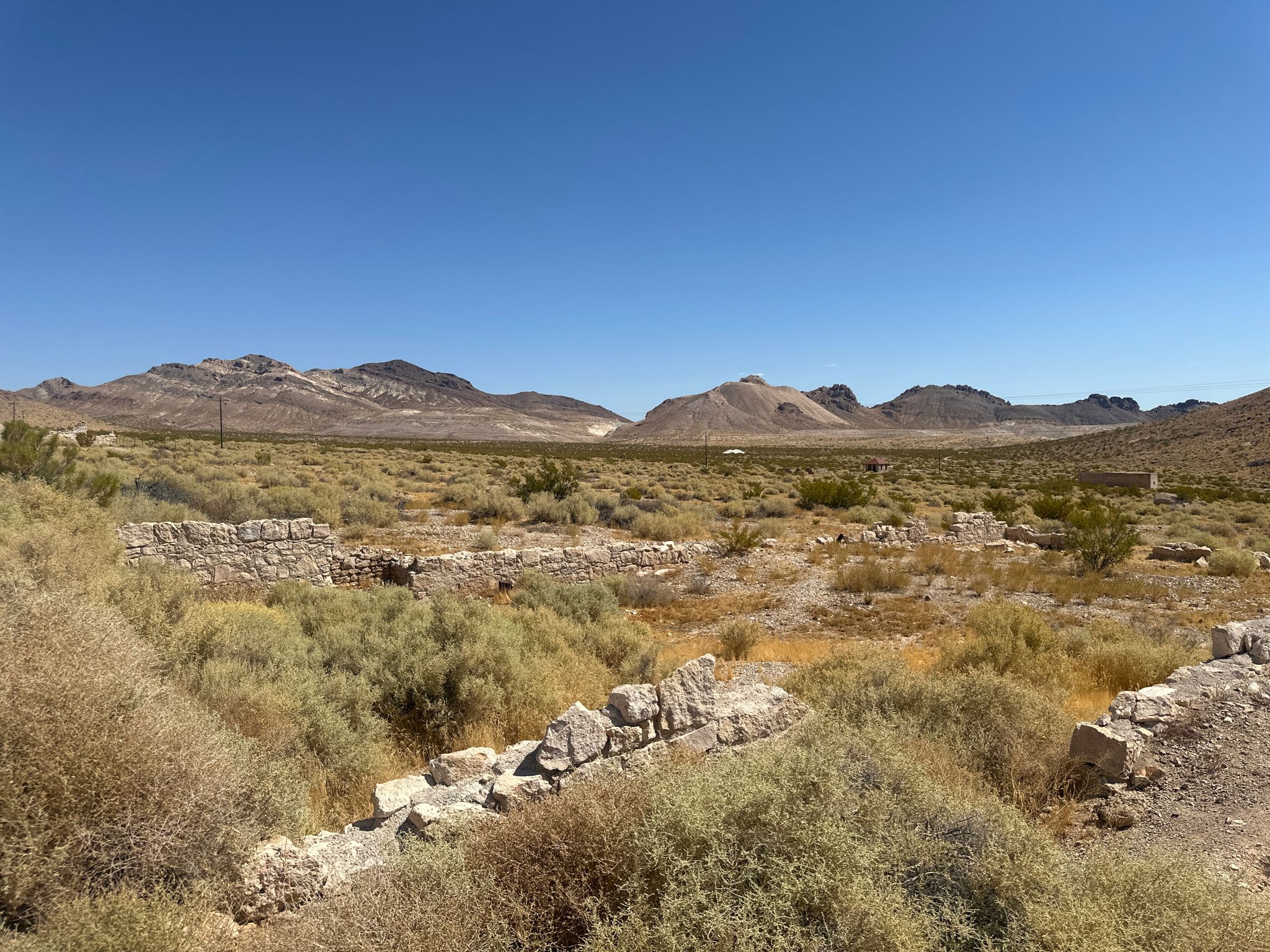 Wide desert landscape with low stone ruins and distant mountains under blue sky. - Rhyolite Ghost Town Trail