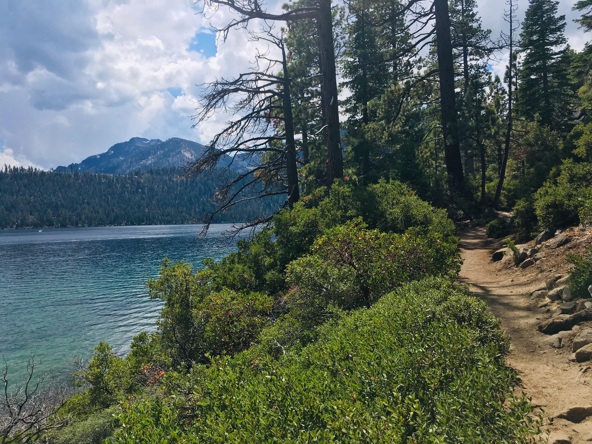 Dirt path beside a clear lake, forested mountains, and cloudy sky. - Emerald Point Trail