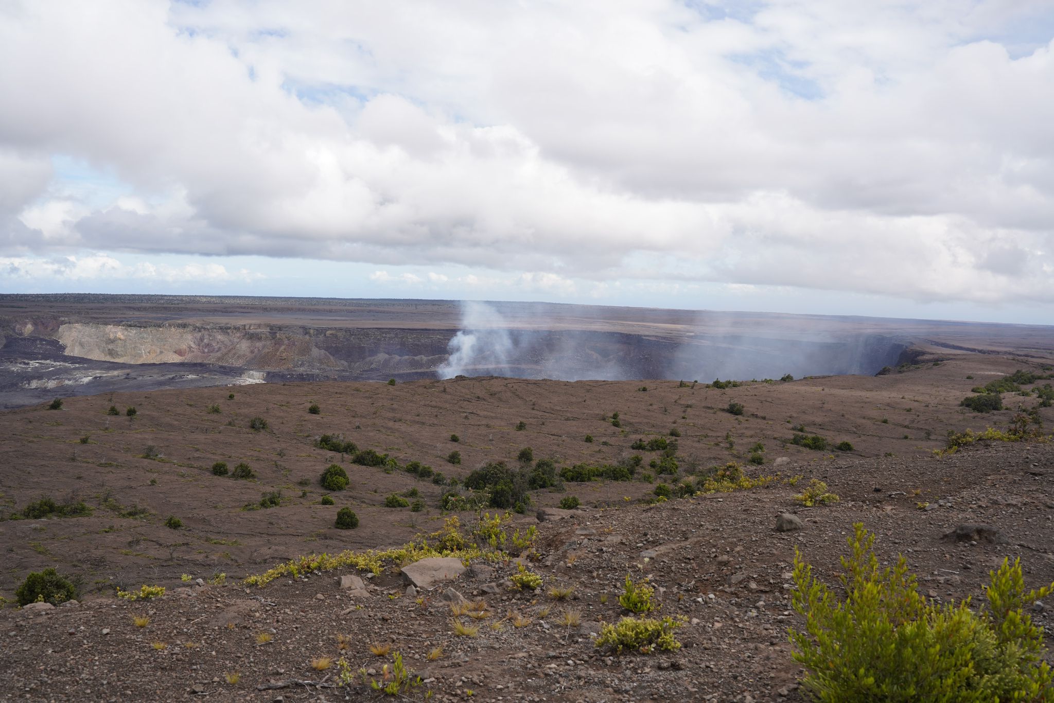 A wide, barren volcanic landscape with a distant steaming crater under clouds. - Crater Rim Trail