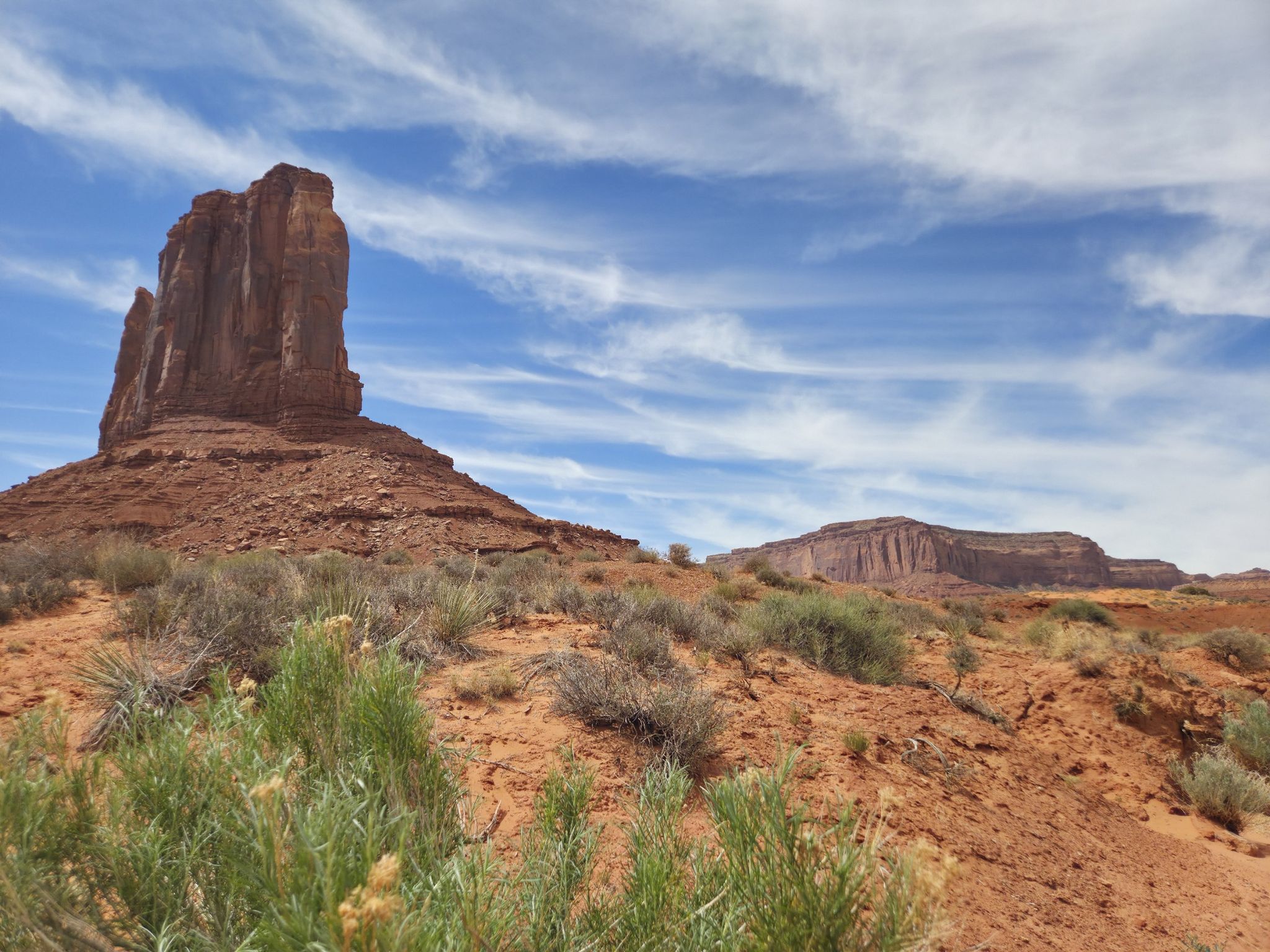 Large red rock formation on left, desert shrubs in foreground, under a blue sky. - Wildcat Trail loop from Monument Valley Navajo Tribal Park