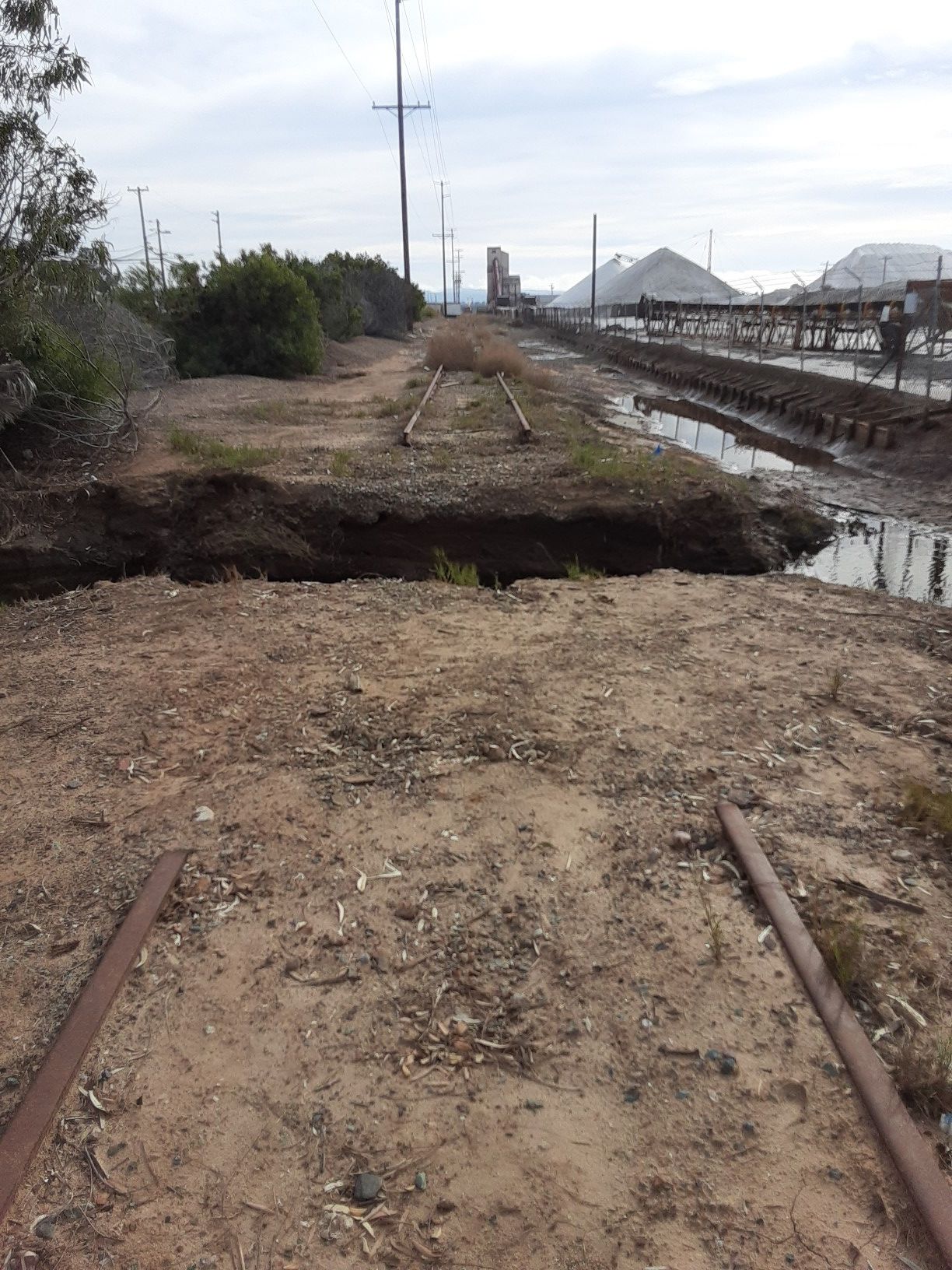 Broken railroad tracks cross a muddy ditch with industrial structures in background. - Sweetwater River – Sweetwater Bikeway loop from Palomar Street