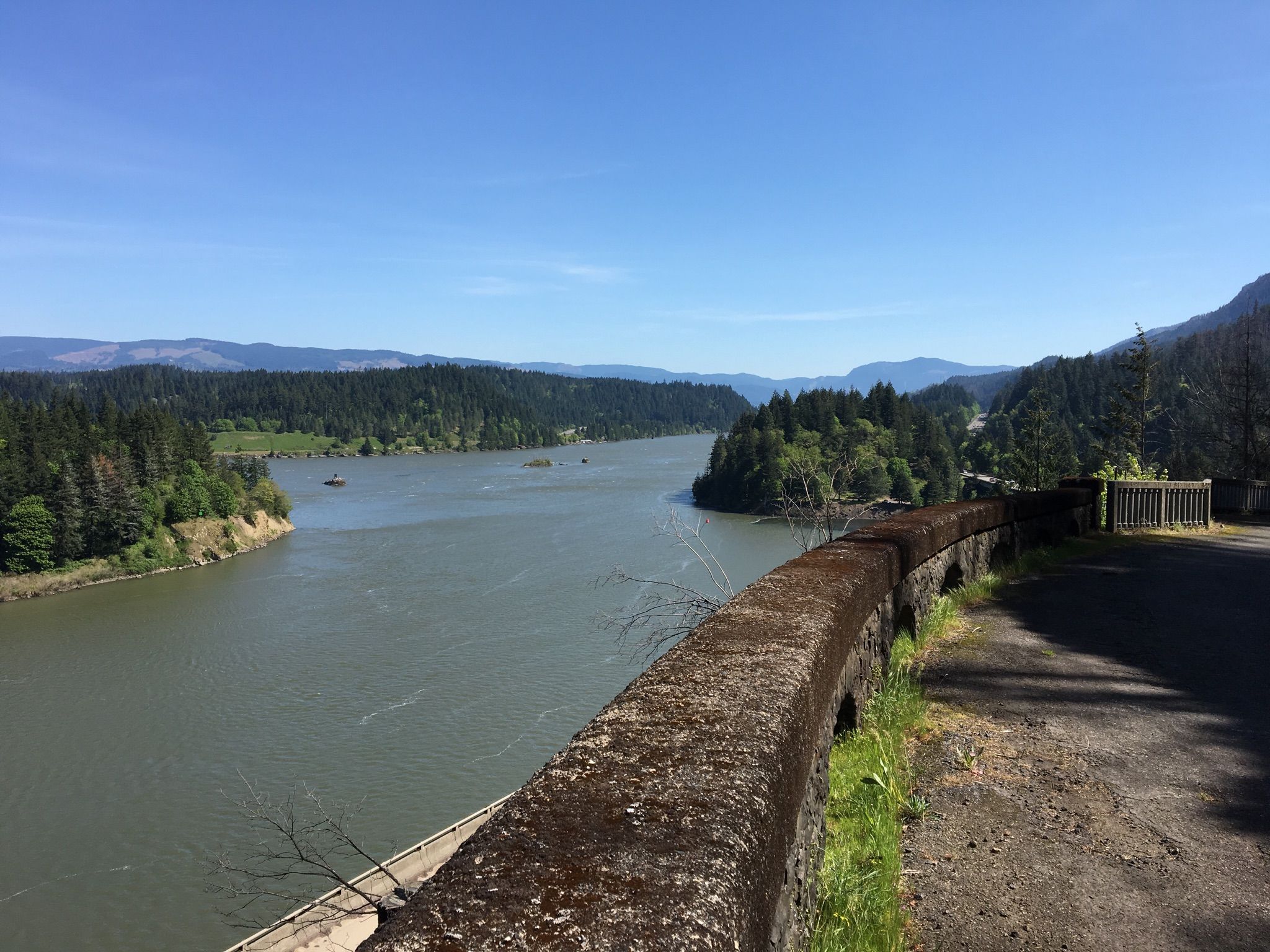 A wide river flows between forested banks, next to a stone wall. - Historic Columbia River Highway State Trail