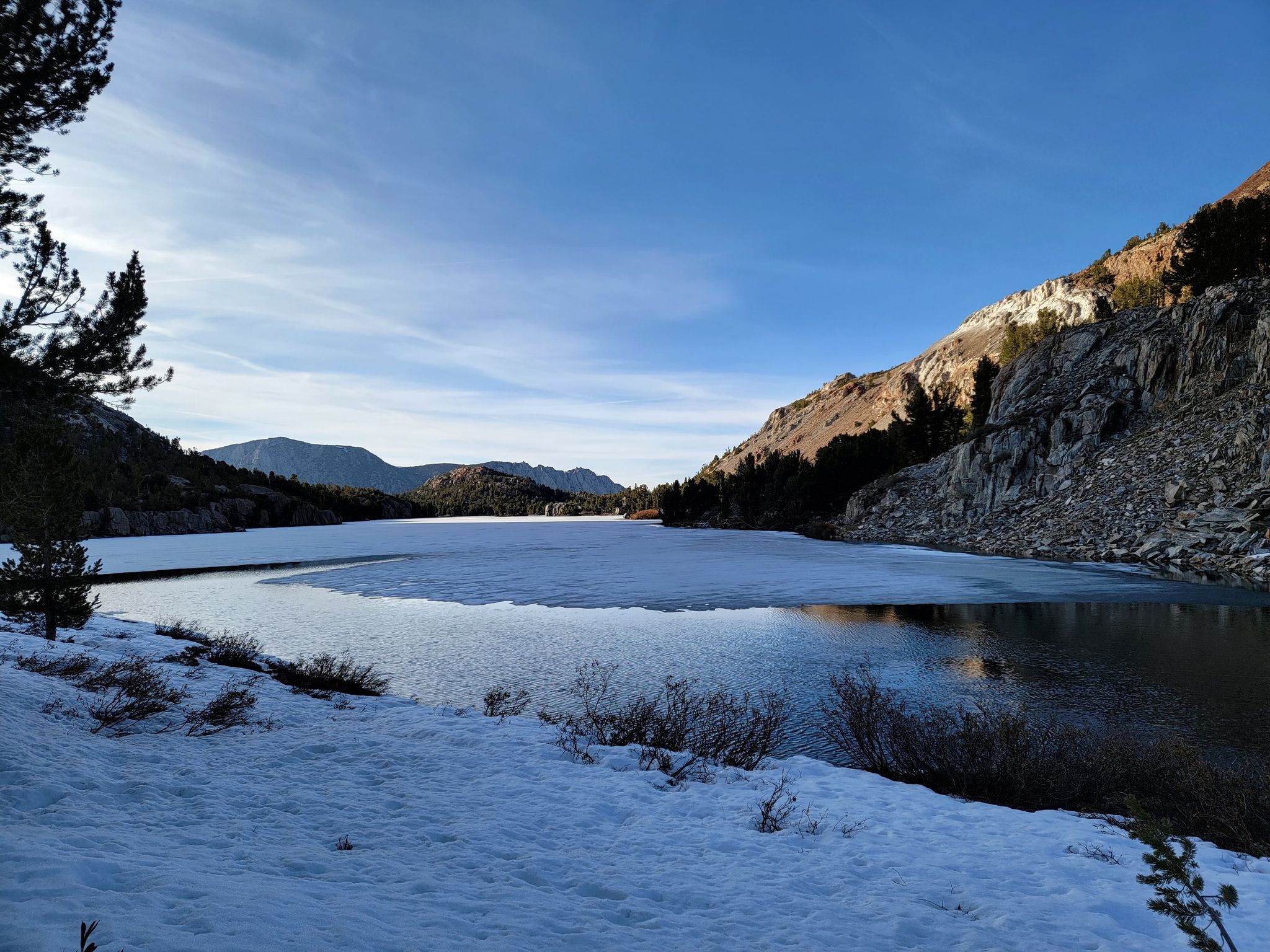 Partially frozen lake surrounded by snowy mountains under a bright sky. - Long Lake, North End – Bishop Pass loop from Hillside Dam