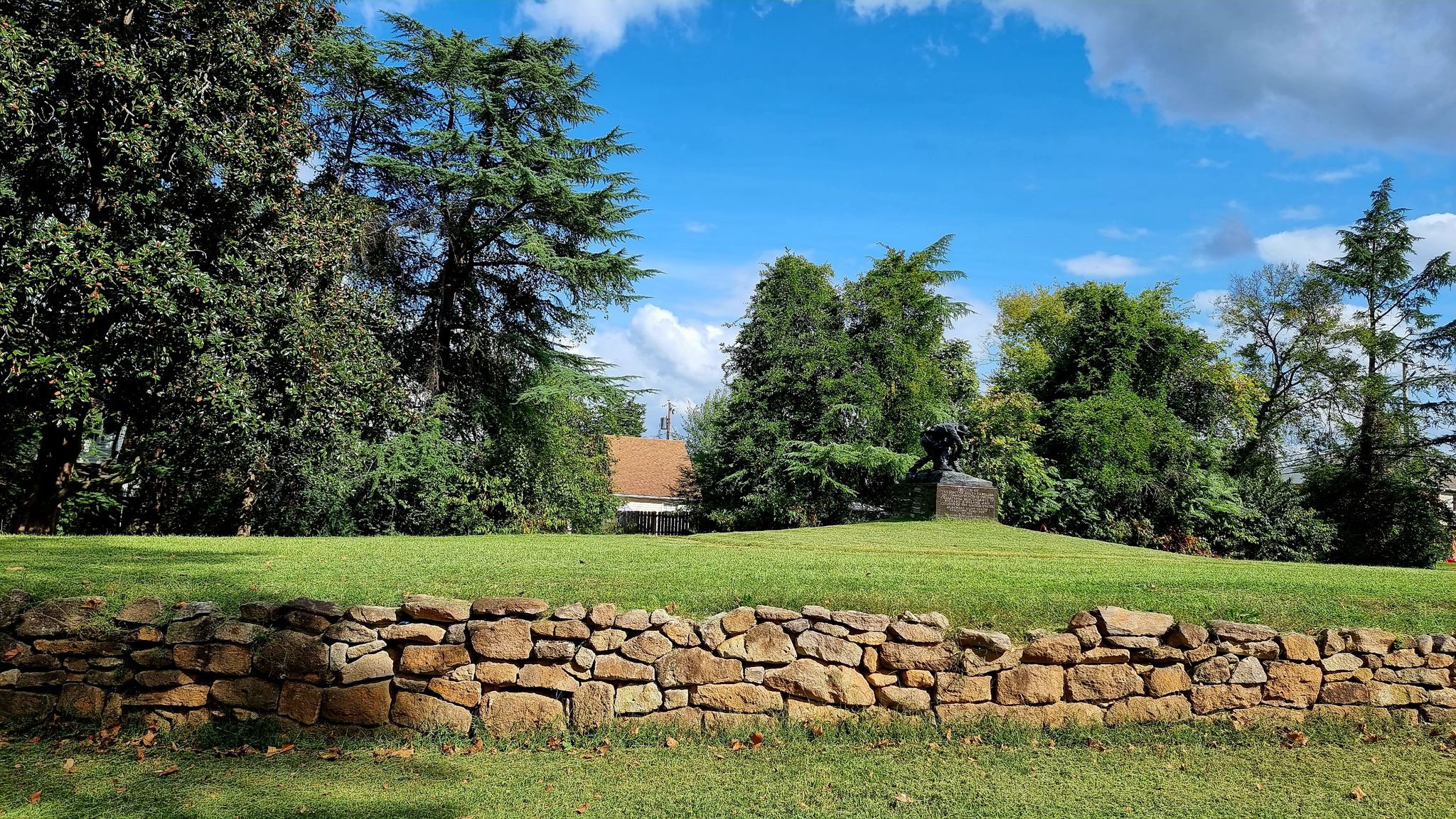 A stone wall in front of a sunny green lawn with dense trees. - Sunken Road Walking Trail