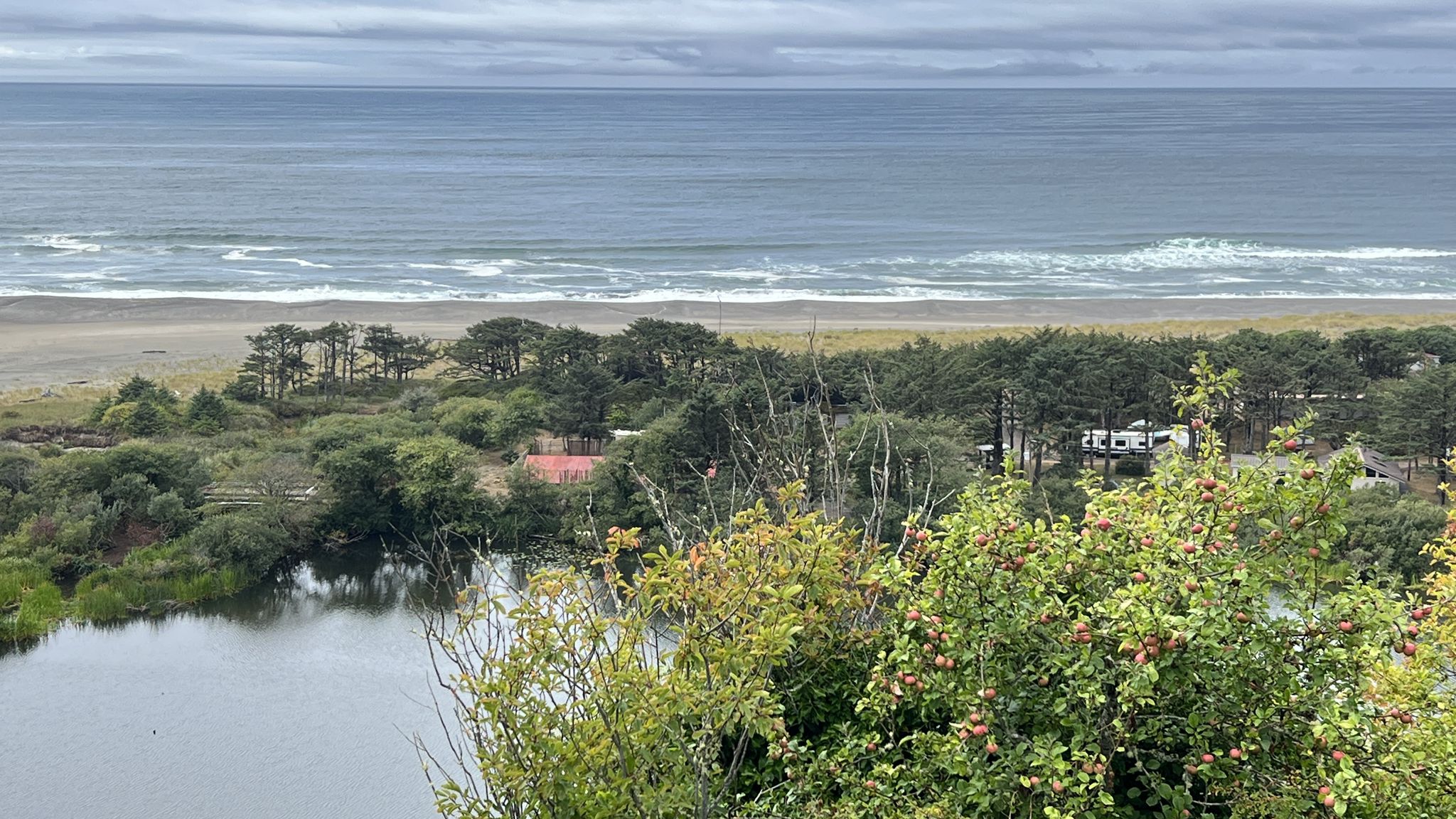 Overcast coastal view of ocean, beach, lake, green bushes, and distant buildings. - Pacific City – Winema Beach Viewpoint loop from Pacific City