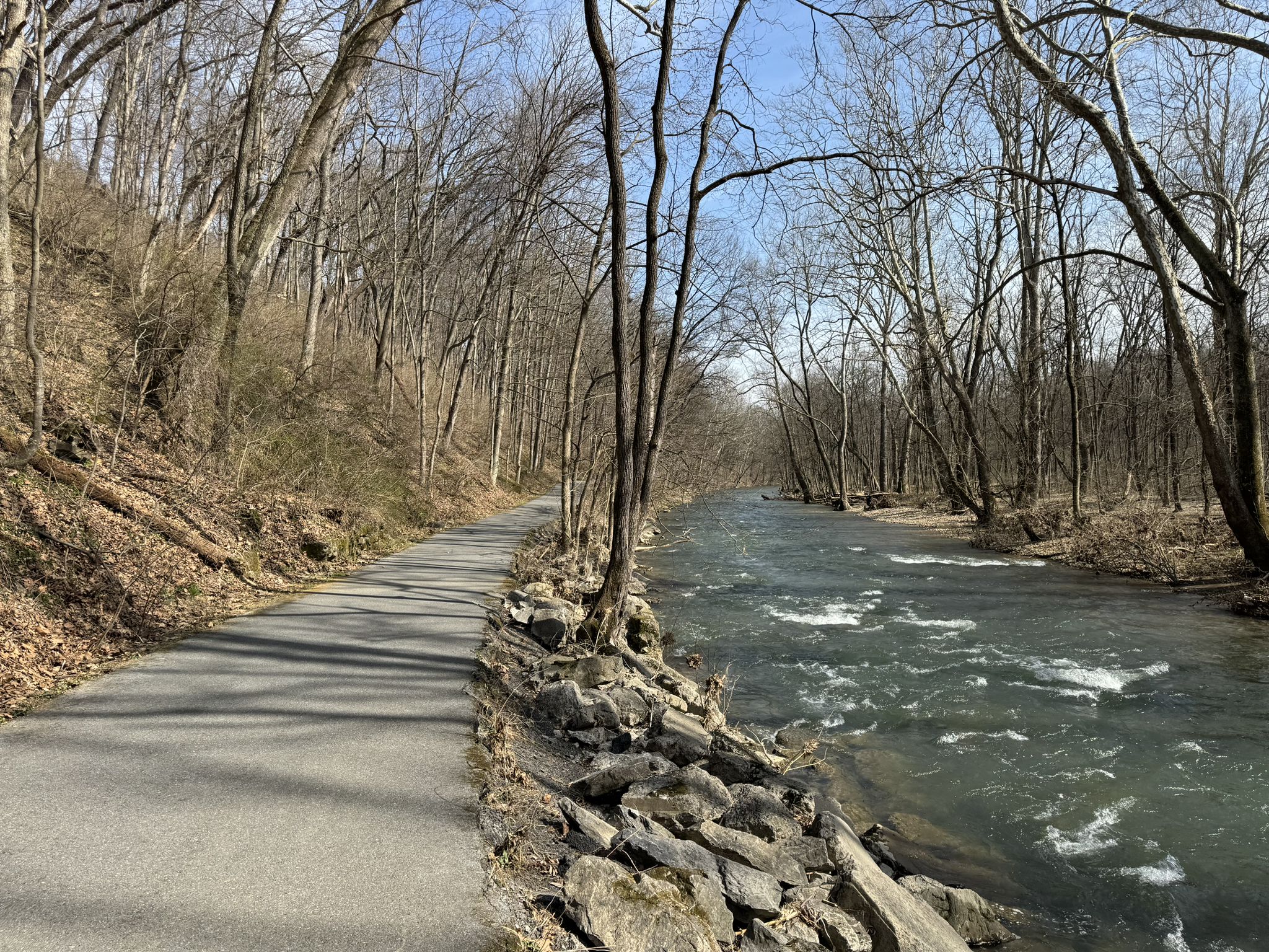 Paved path alongside a rocky river, surrounded by bare trees. - Jordan Creek Greenway