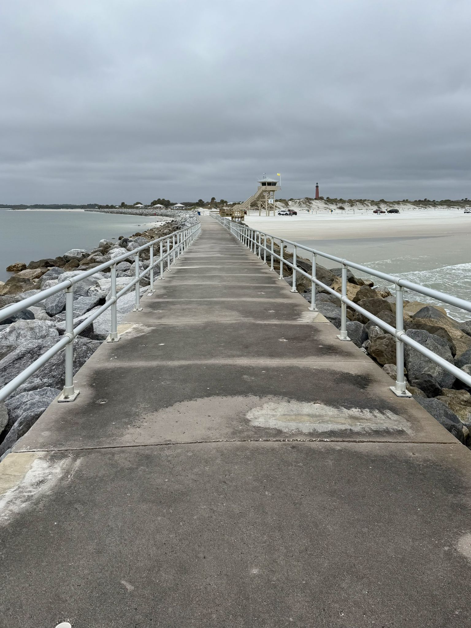 Long concrete pier with railings extends between ocean and sandy beach, overcast. - Ponce Inlet Jetty Trail