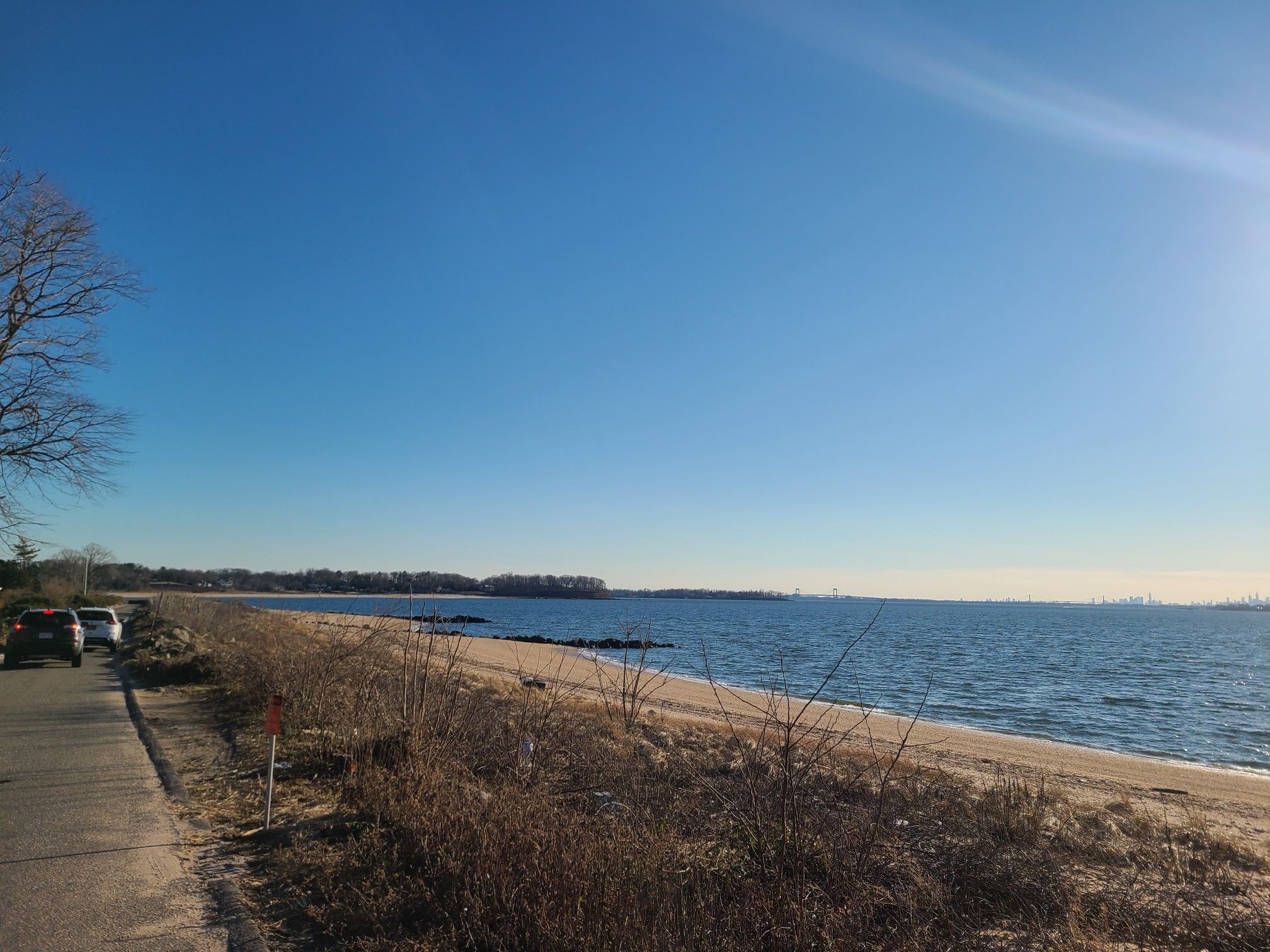 Sunny beach with calm blue ocean, clear sky, and cars on a nearby road. - Sands Point - Lighthouse Road – Port Washington Waterfront loop from Port Washington