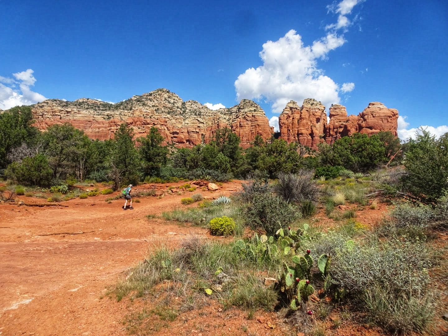 A person walks on a red dirt path towards distant red rock cliffs. - Thunder Mountain – MTB Trails in Sedona