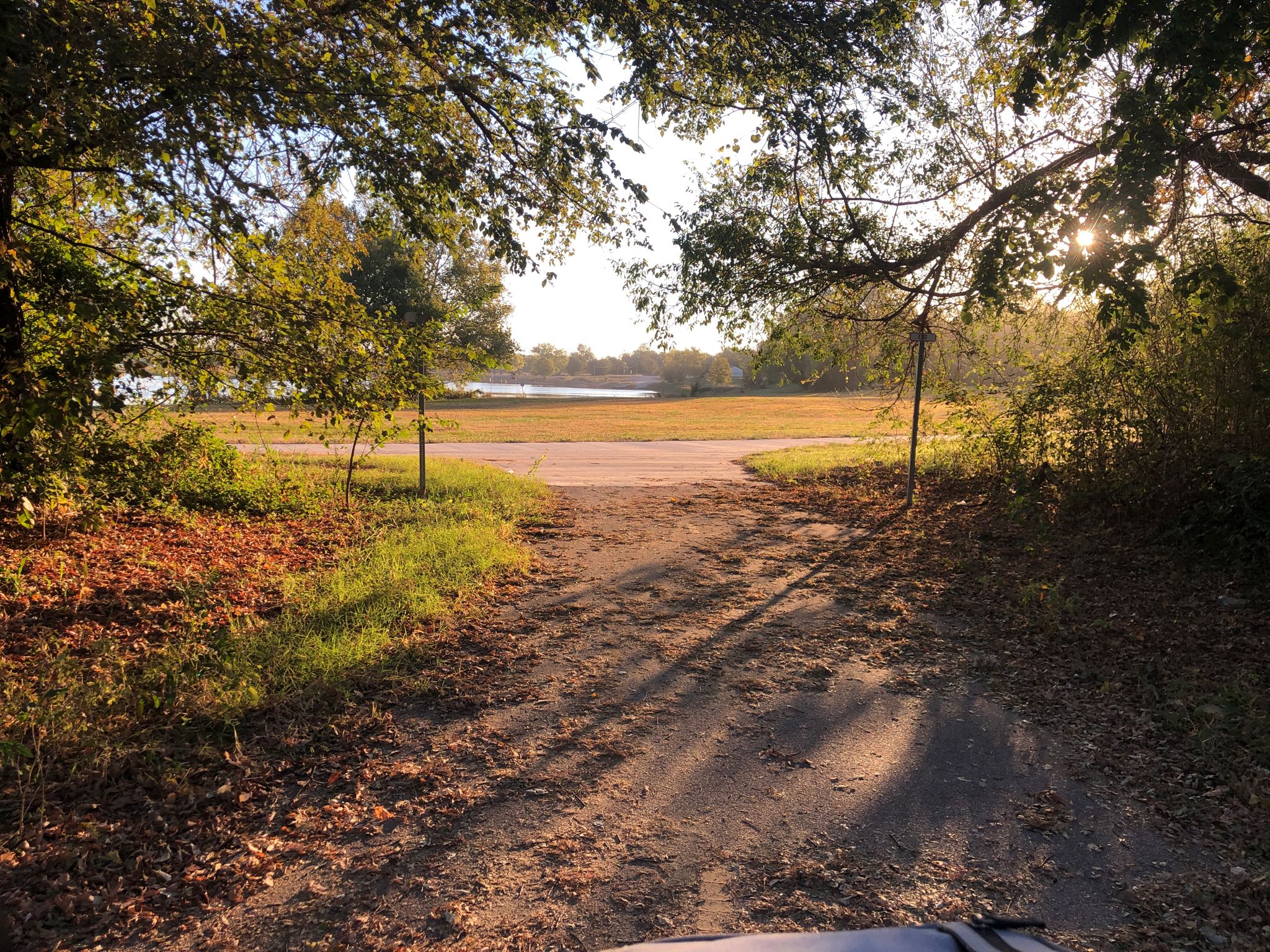 Sunlit dirt path framed by trees, leading to an open field and water. - Eagle Lake Trail