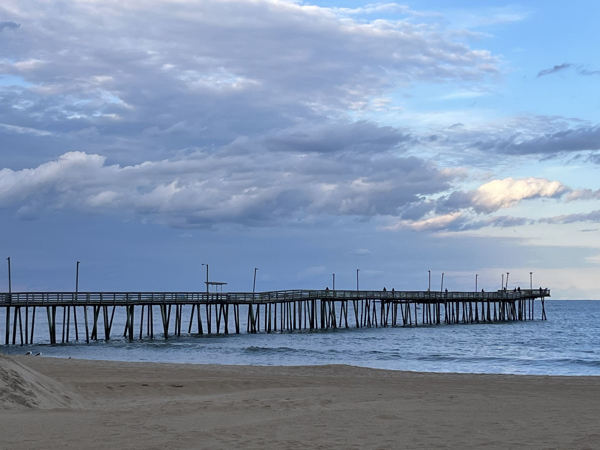 Long wooden pier extending into the ocean from a sandy beach under cloudy skies. - Virginia Beach Boardwalk