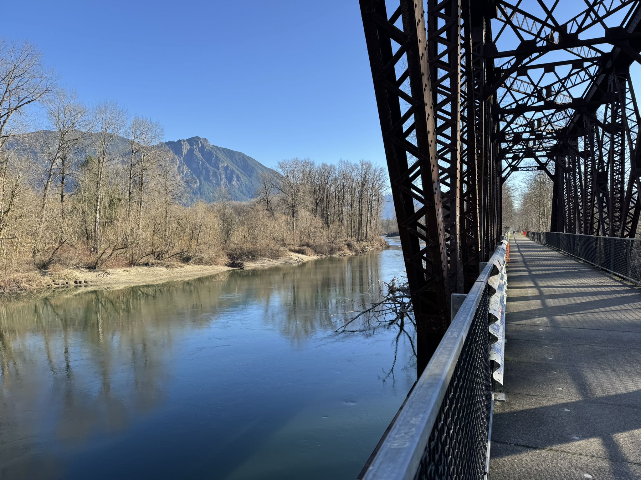 Metal bridge spanning a calm river with bare trees and mountains. - Snoqualmie Valley Trail to Three Forks Loop