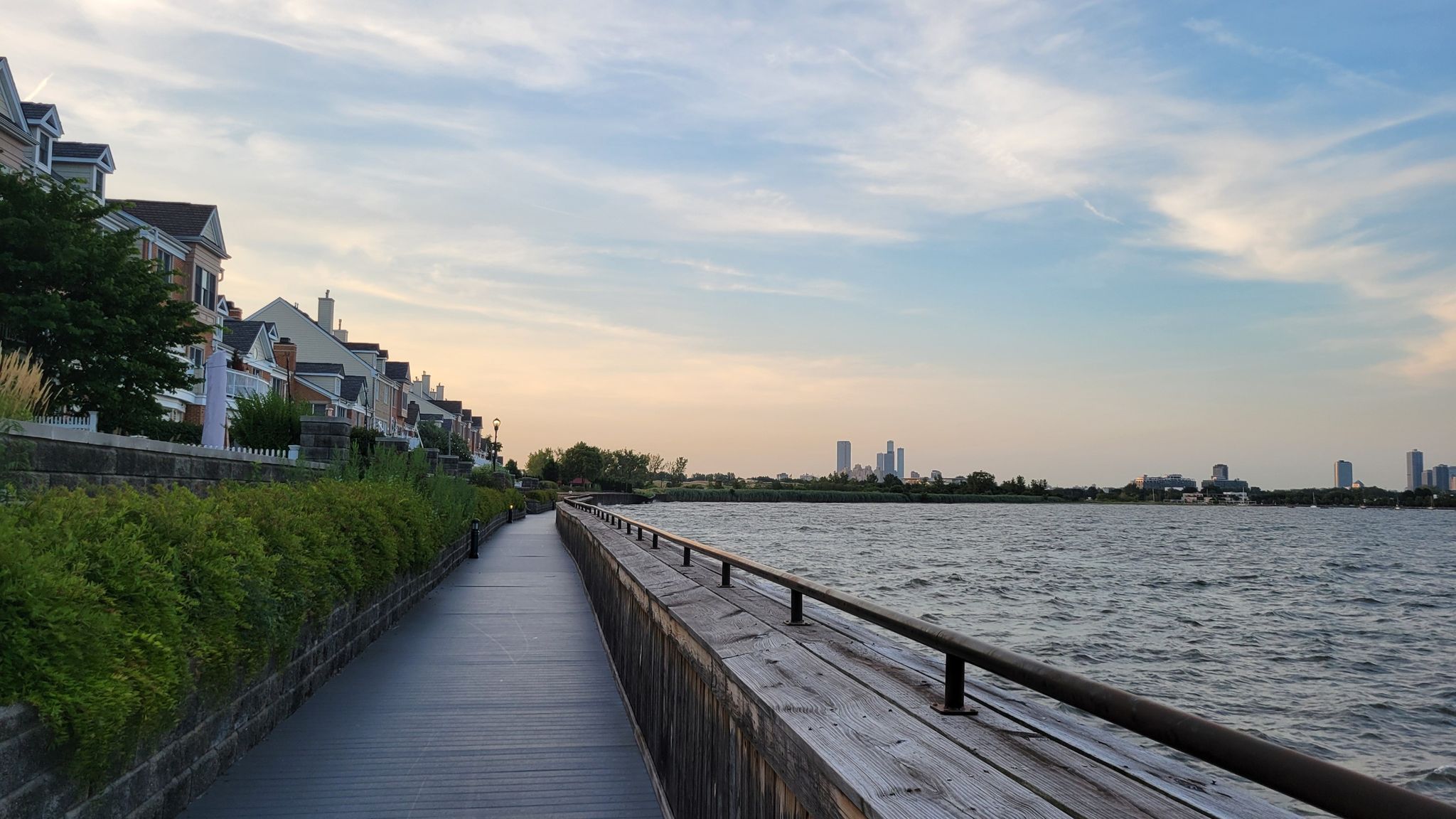 A boardwalk beside water with houses and a distant city at dusk. - Caven Point Trail