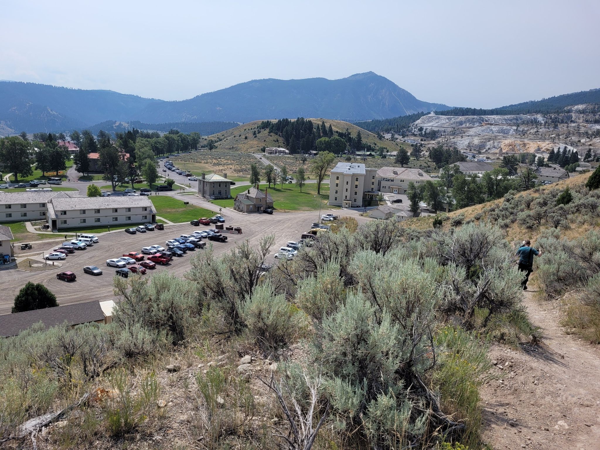 Overlook of a town with buildings and mountains under a cloudy sky. A person walks on a trail. - Beaver Ponds Loop Trail