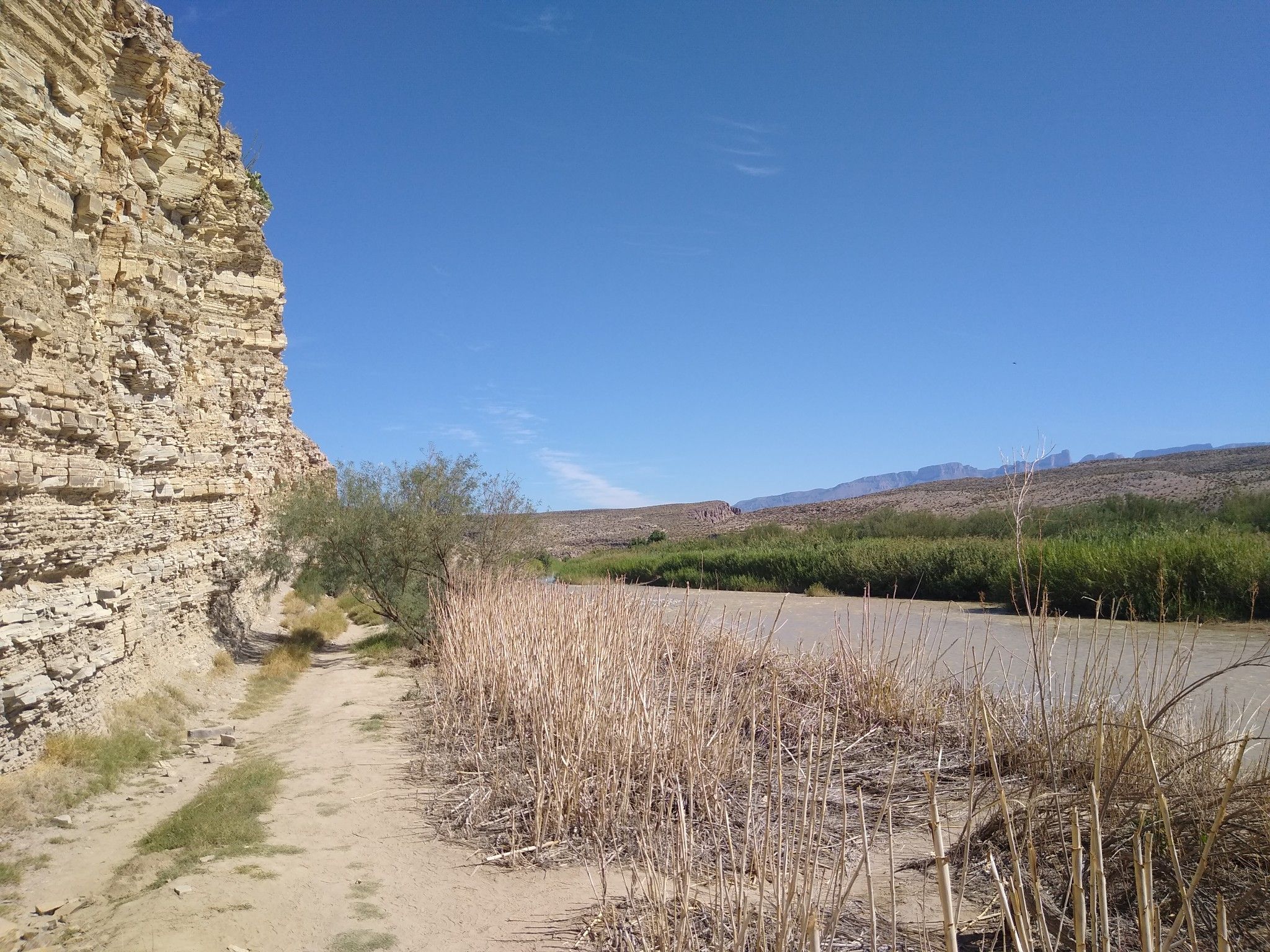 Dirt path next to a tall rock cliff and a river with dry reeds. - Old Bathhouse Stone Crossing – Big Bend Hot Springs loop from Hot Springs