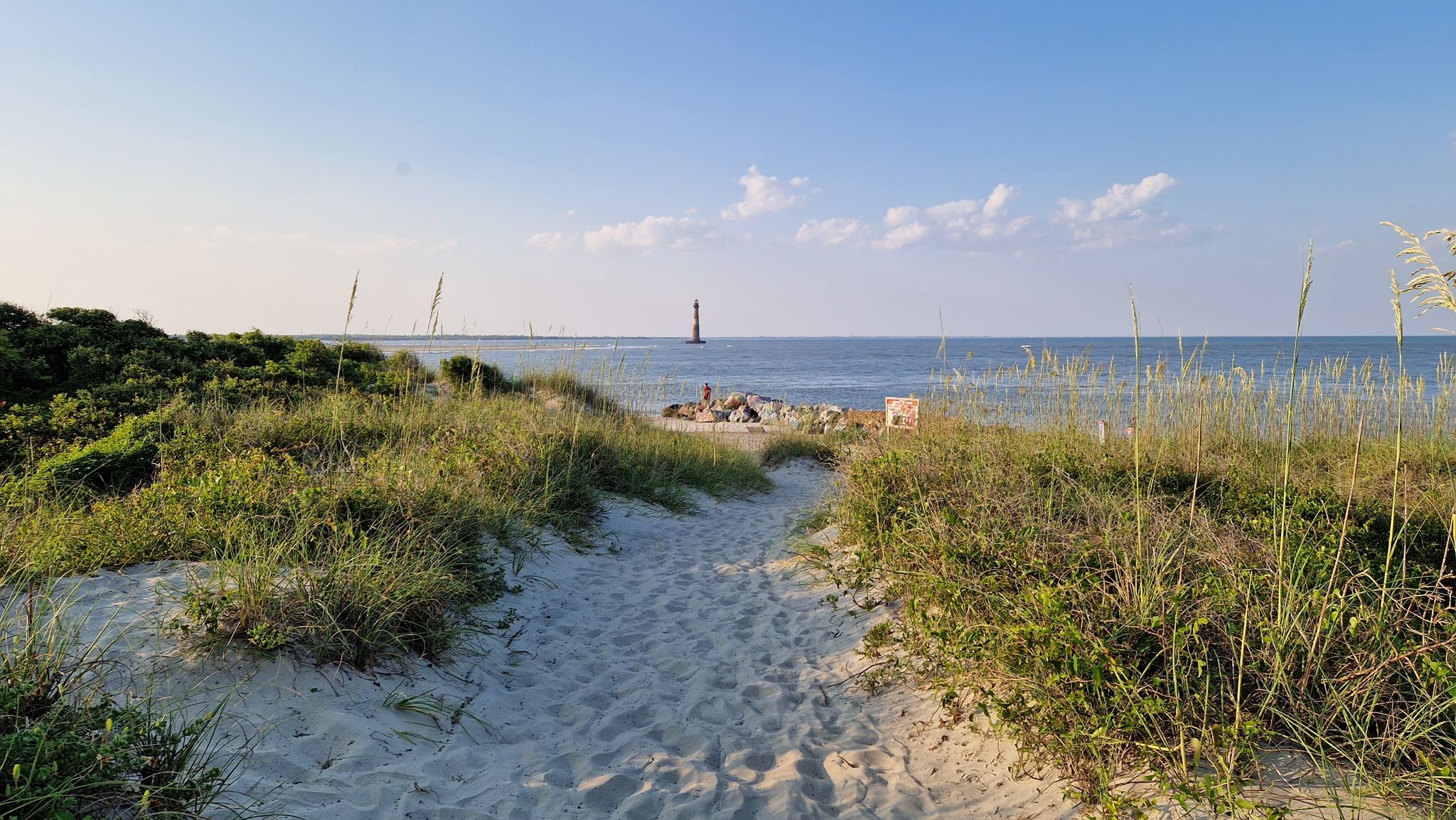 Sandy path leads to a sunny beach with ocean, dunes, and distant lighthouse. - Morris Island Lighthouse Trail