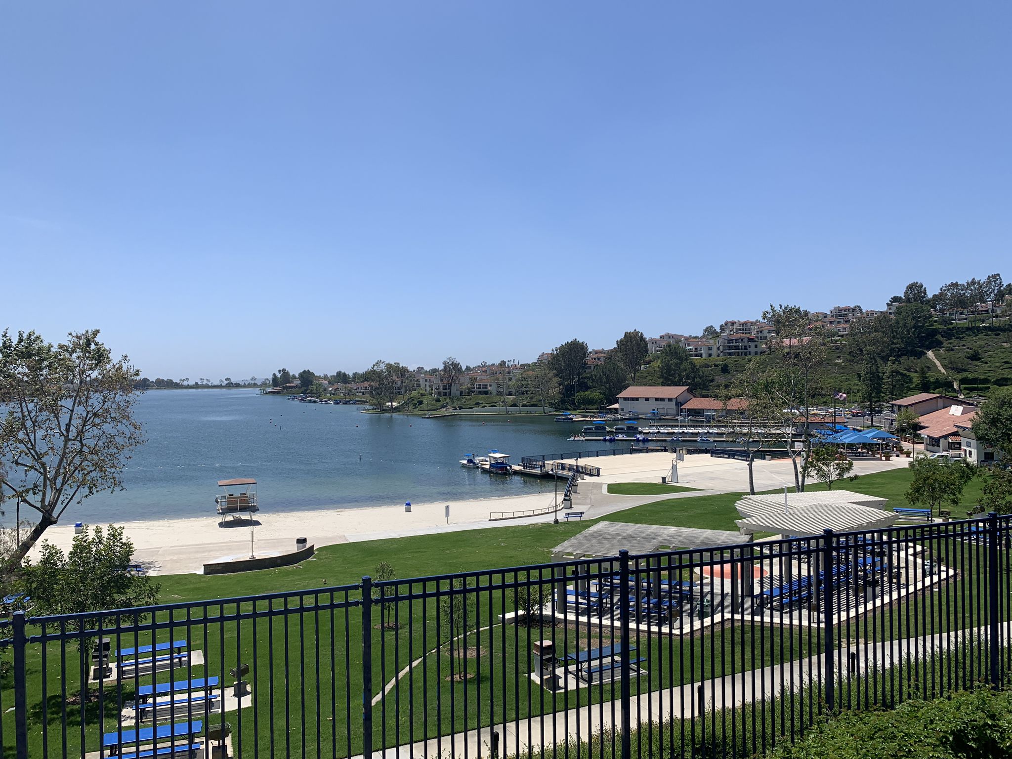 Bright daylight view of a lake with a sandy beach, dock, and park. - Lake Mission Viejo Loop