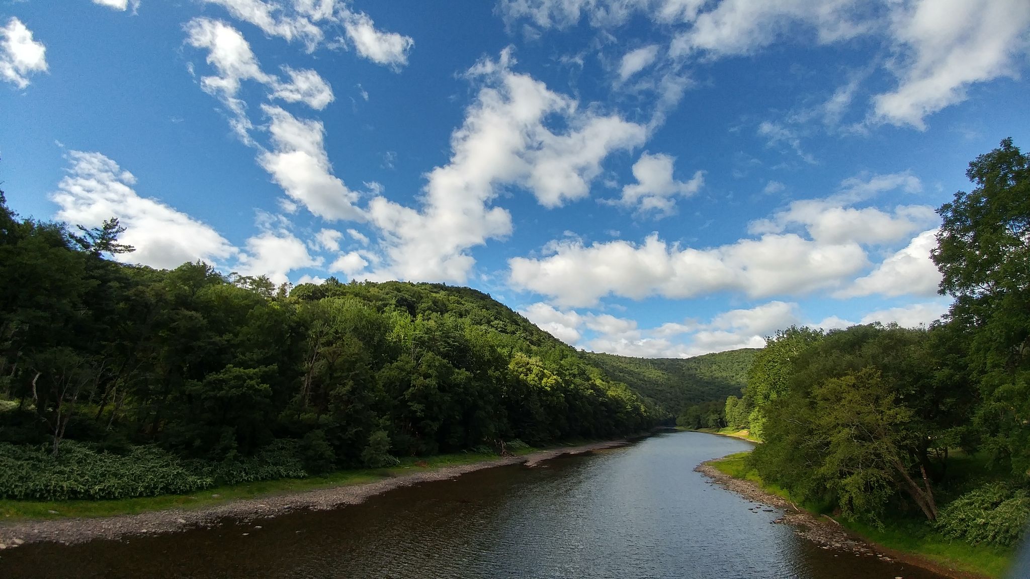 Wide river flowing between lush green forested hills under a bright blue sky. - Little Pine Lake – Vista loop from Little Pine State Park