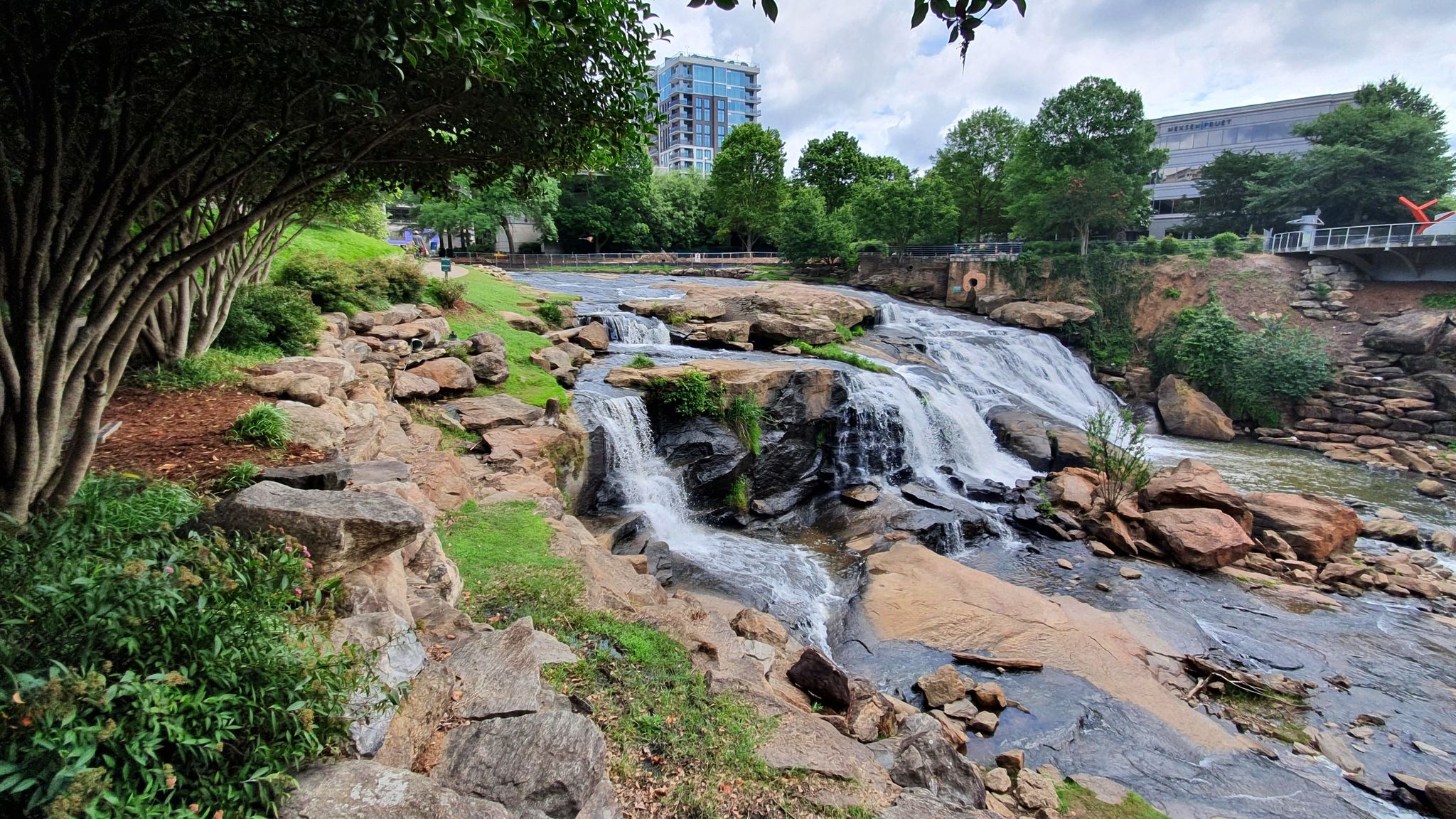 Cascading waterfall over rocks, lush greenery, and distant city buildings under cloudy sky. - Paris Mountain and Swamp Rabbit Trail Loop