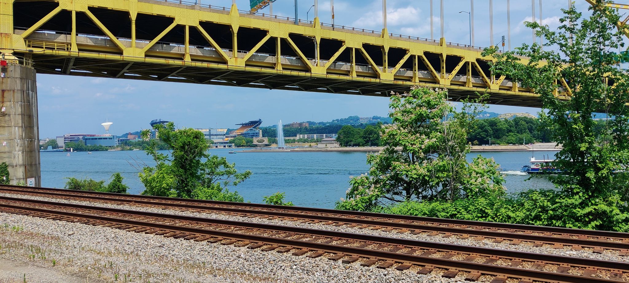 A yellow truss bridge spans over railroad tracks and a river with a boat. - Station Square Trail