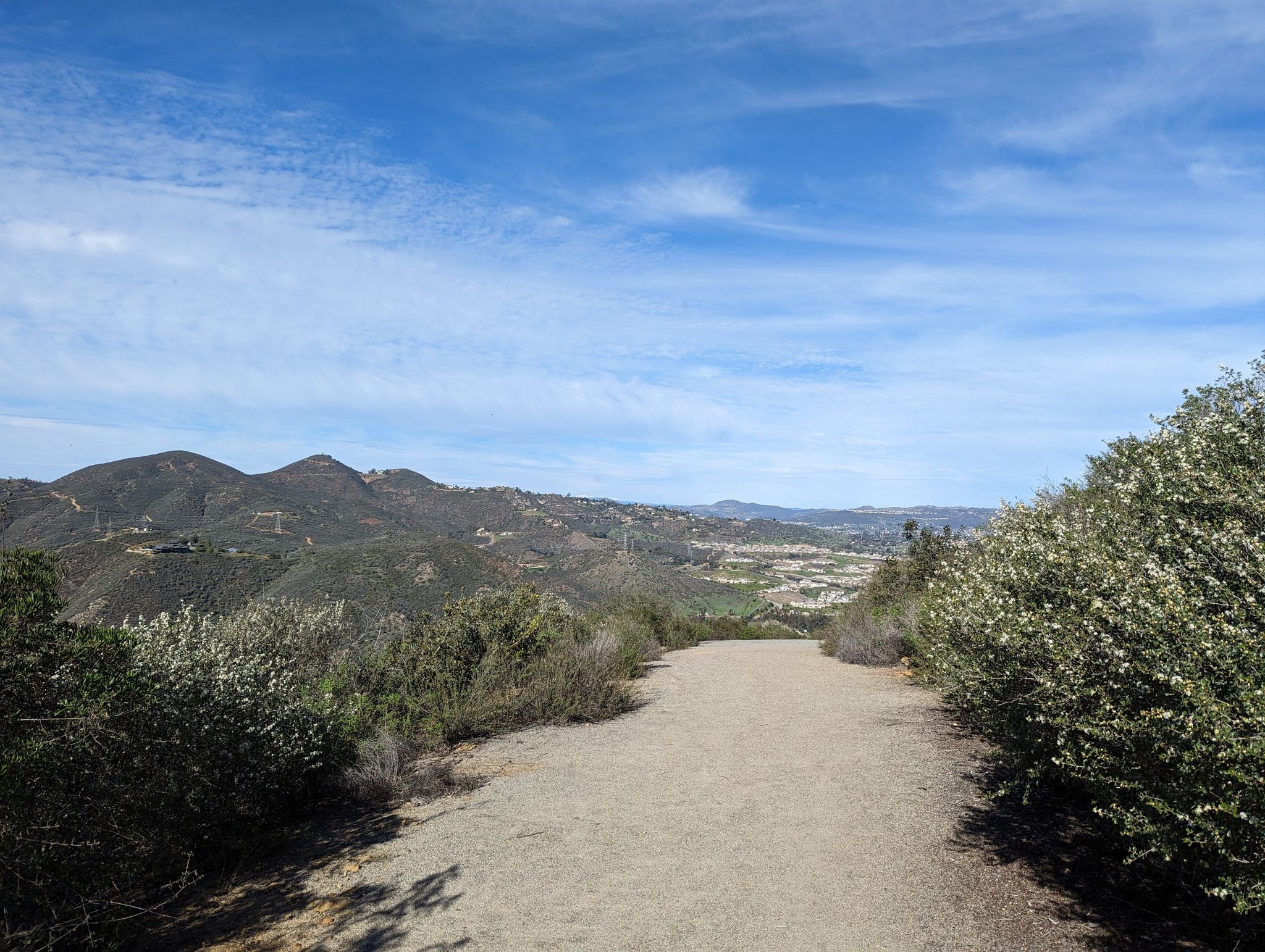 Dirt path descends into a valley with green hills under a bright sky. - Lake Hodges Overlook Trail