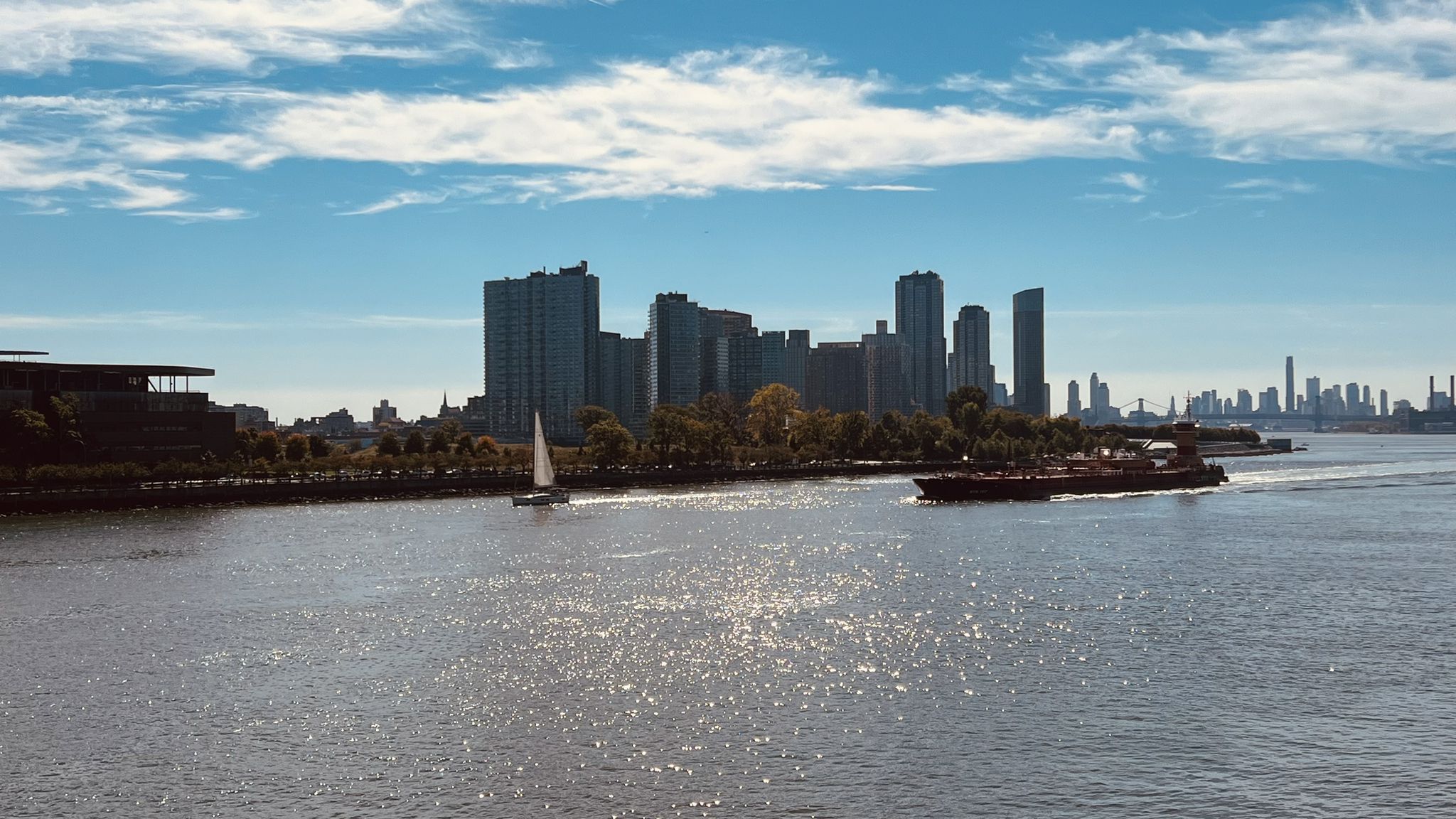 Wide river with a sailboat and barge, distant city skyline under blue sky. - East River and Randalls Island Greenway Loop