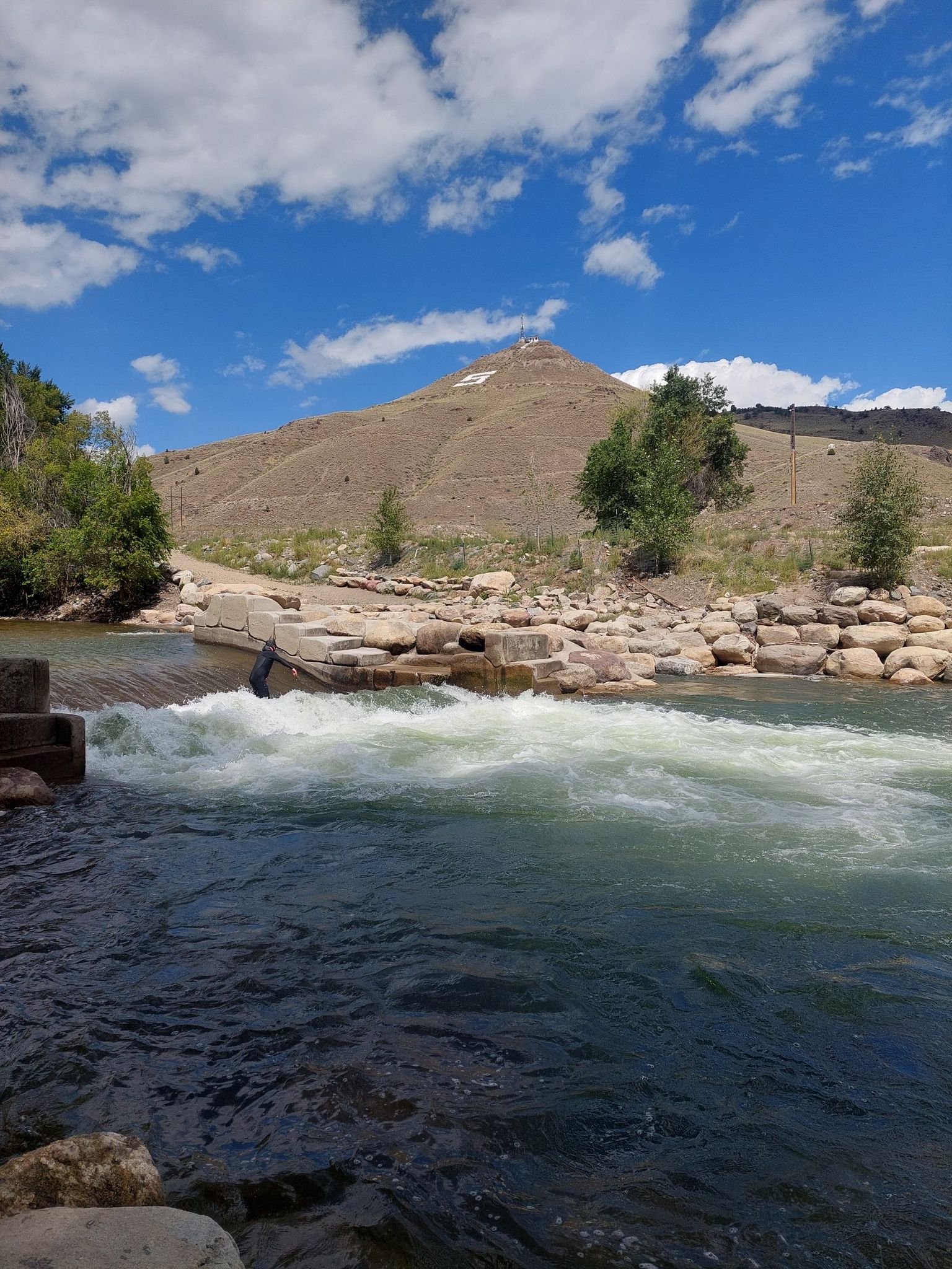 Rushing river water with rocky banks, leading to a barren hill under blue sky. - Riverside Park – Along the Arkansas River loop from Salida