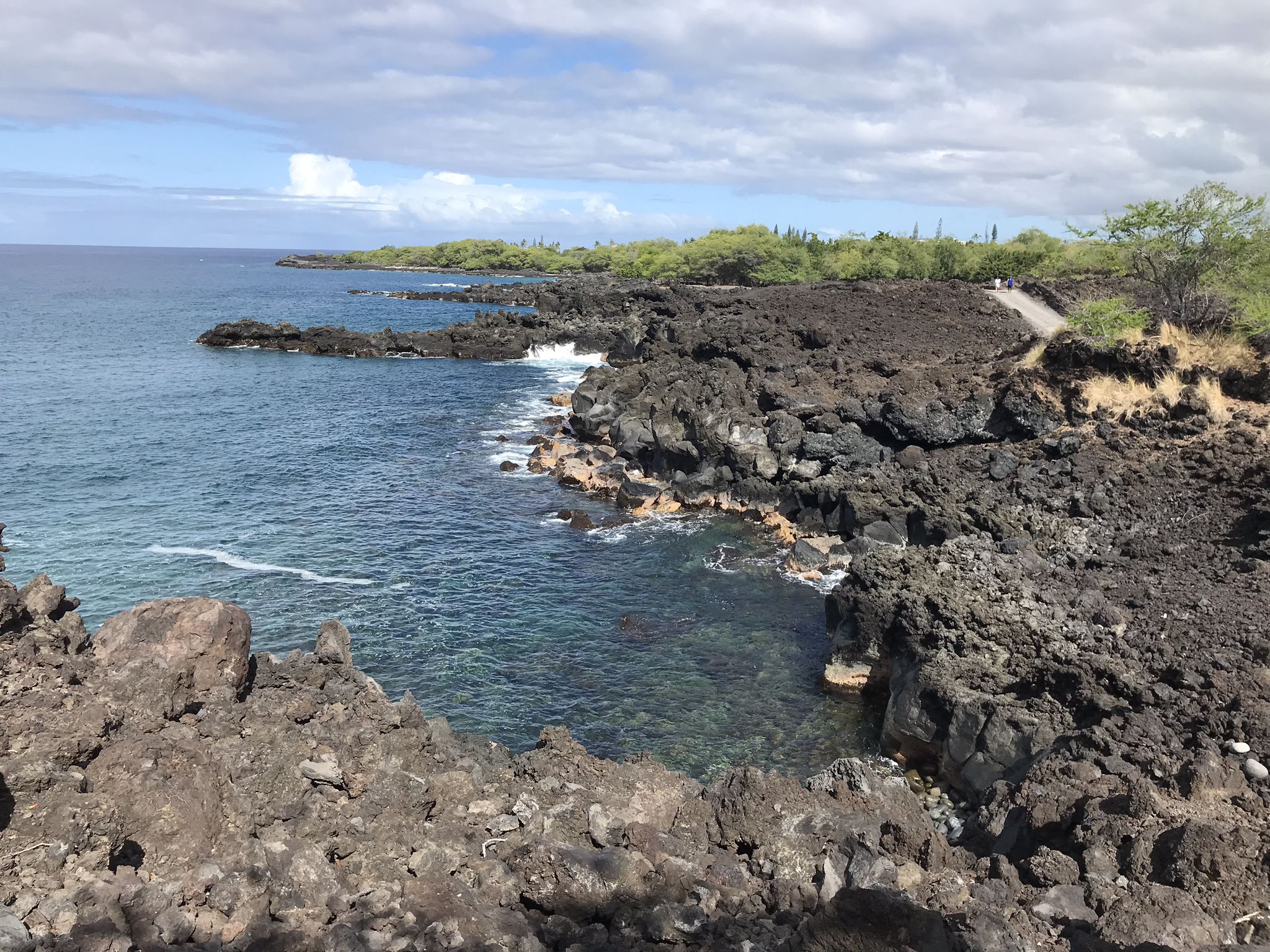 Rocky volcanic coastline with clear blue ocean under a partly cloudy sky. - End of the World Trail