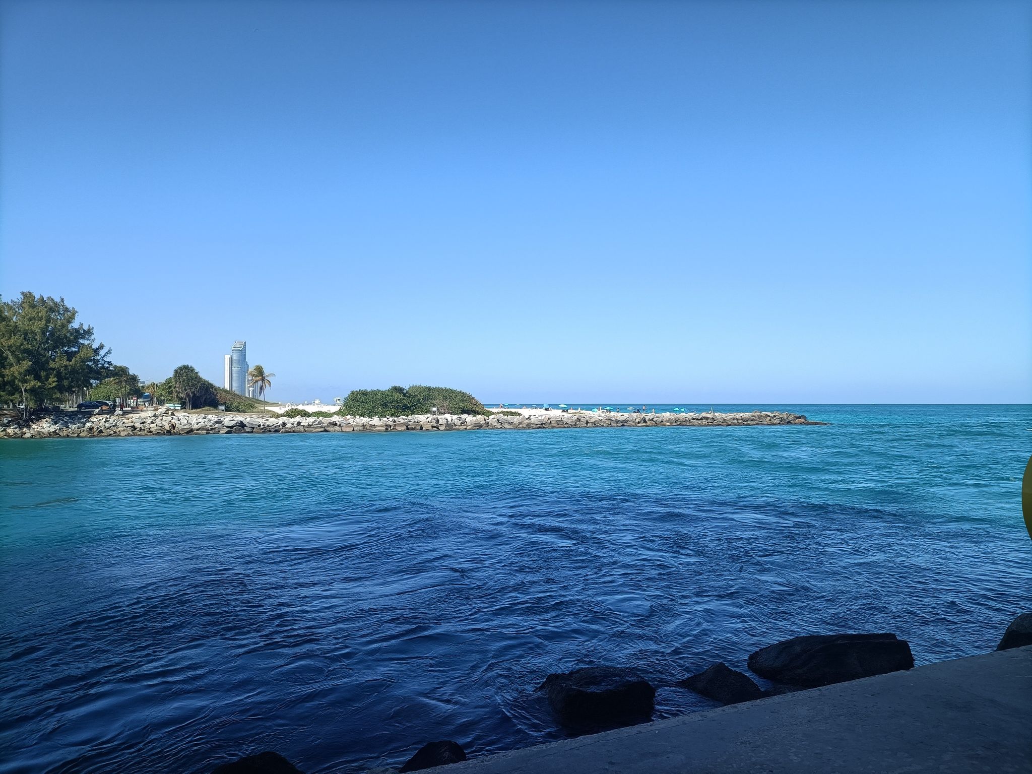 Calm blue water with a distant rocky shore and city building. - Rotraut Color Sculptures loop from Surfside