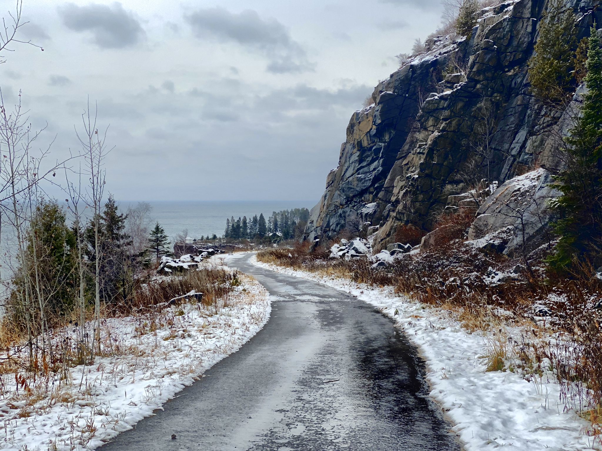 Icy paved road winding along a rocky cliffside with snow and distant water. - Gitchi-Gami State Trail
