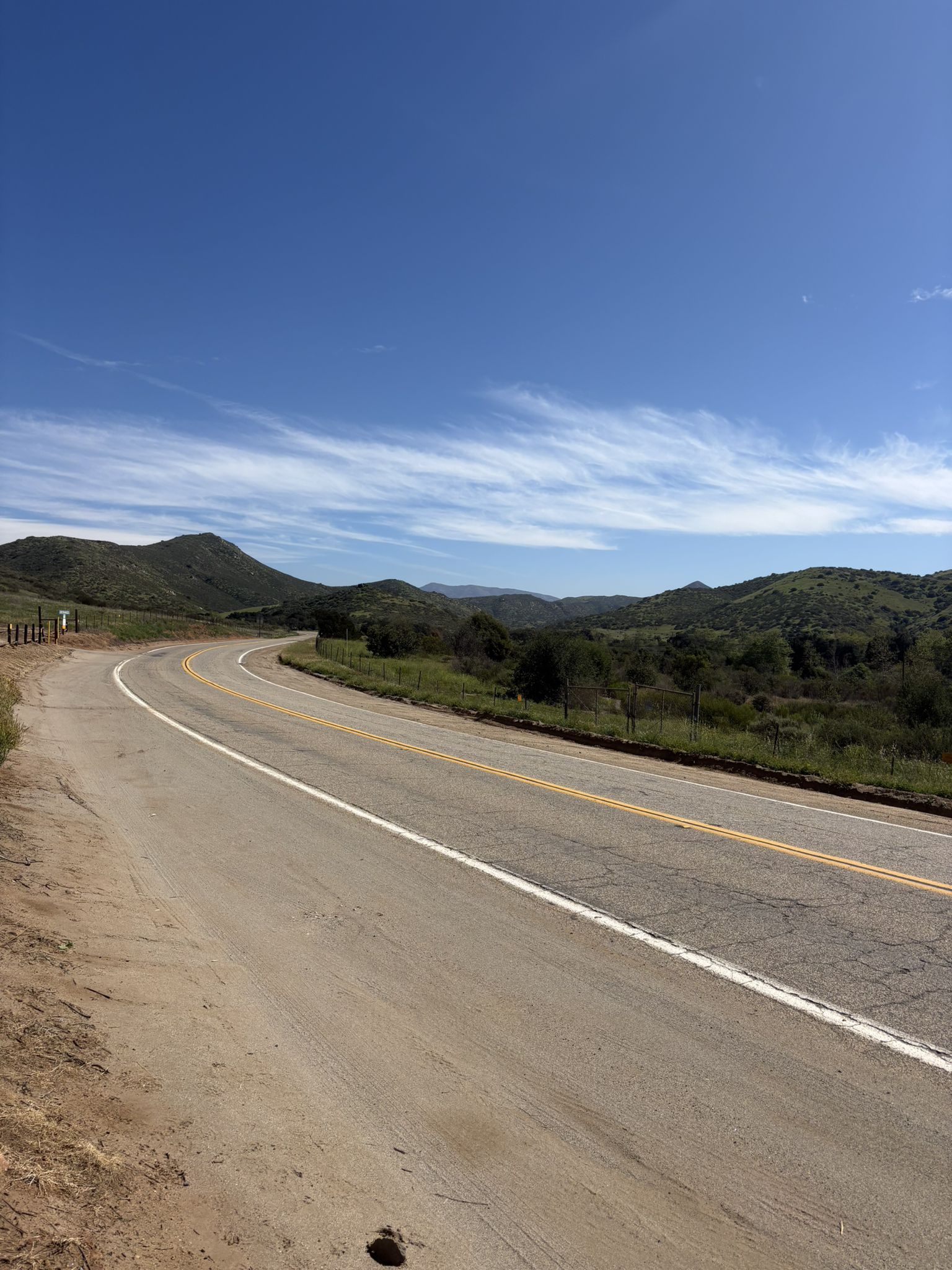 A winding road curves through green hills under a bright blue sky. - Daley Ranch Meadow Trail