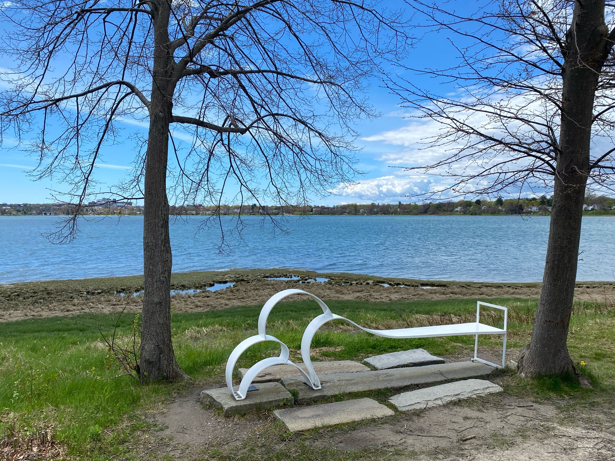 A modern white bench sits between two bare trees by a lake under a blue sky. - East Coast Greenway: Back Cove to Pine Grove Preserve