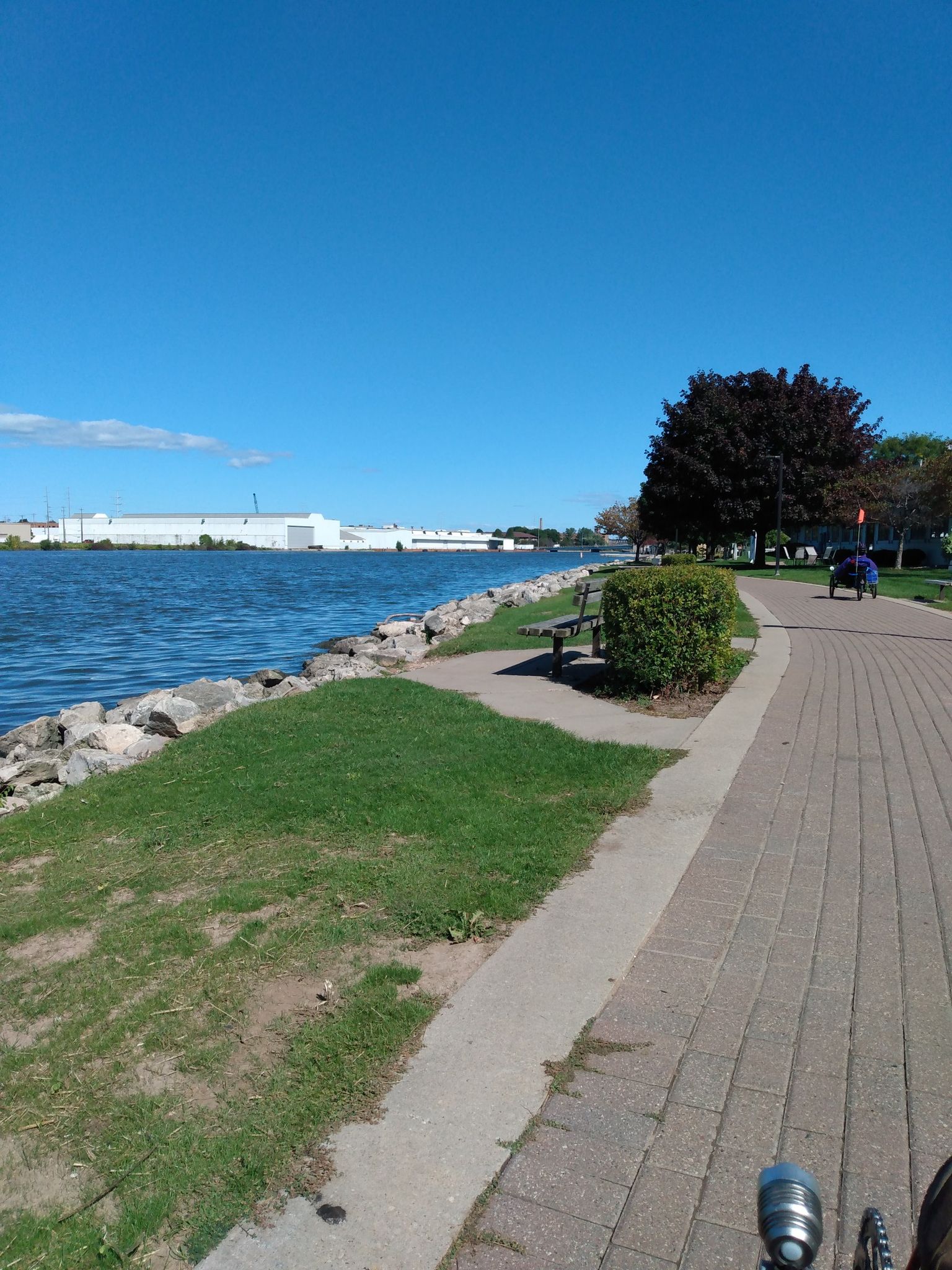 Sunny day on a paved path beside blue water, with a cyclist in distance. - Fox River Trail – Downtown Green Bay loop from De Pere