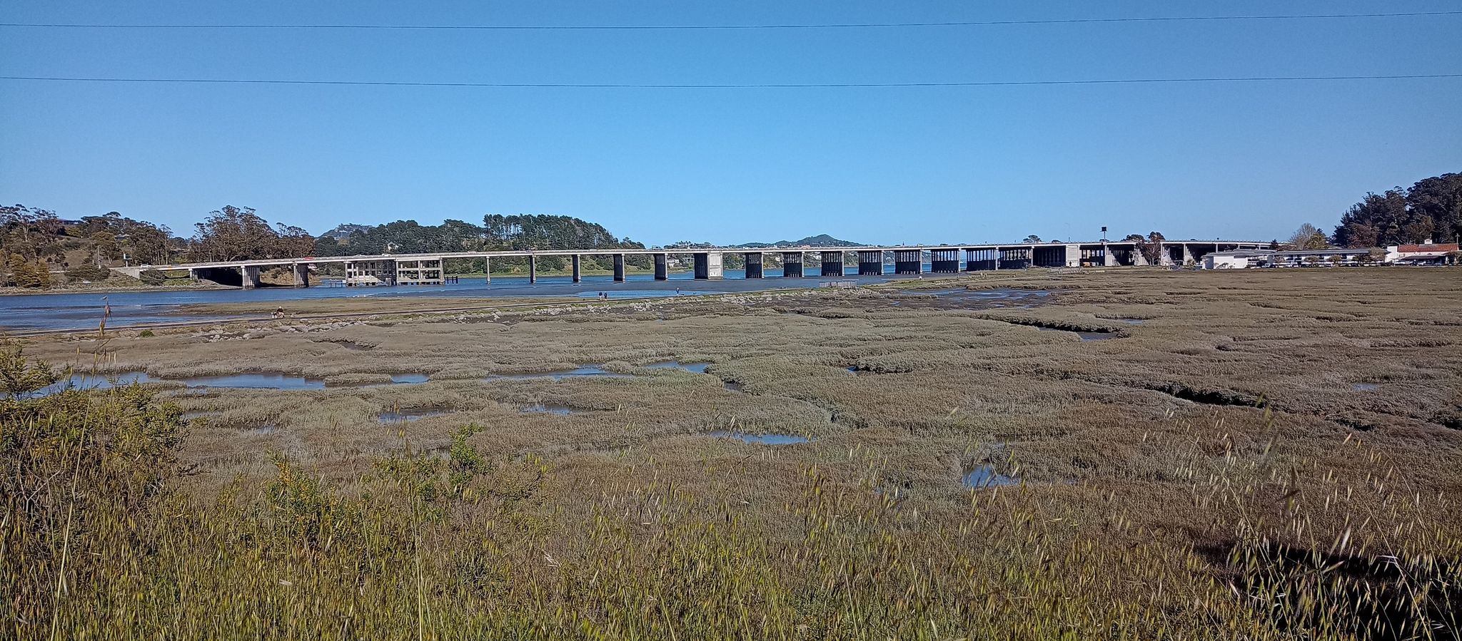 A long bridge spans calm water and expansive marshland under a clear sky. - Muir Beach Overlook – Shoreline Highway in Mount Tamalpais State Park loop from Tamalpais-Homestead Valley