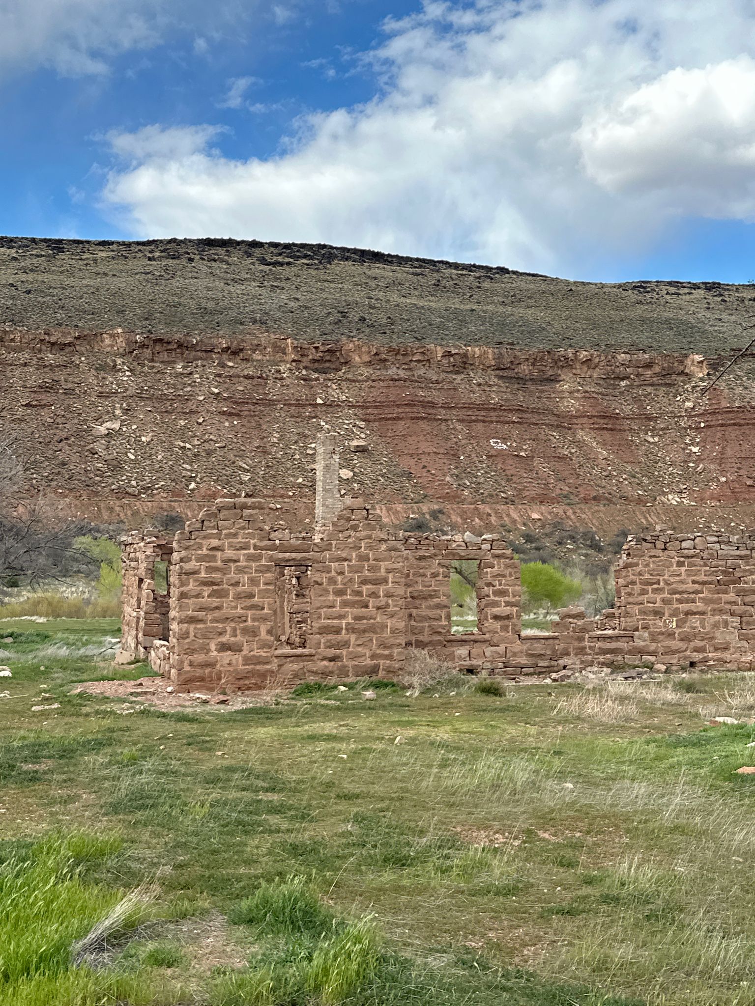 Stone ruins stand in a green field before a large red rock cliff. - Veyo loop