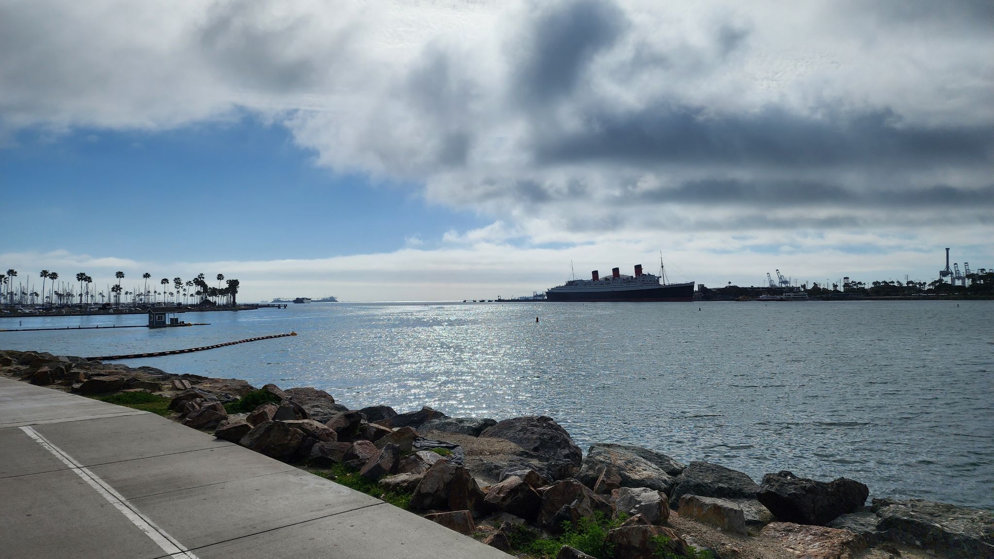 Harbor view with a large ship, palm trees, and rocky shore under cloudy sky. - Long Beach Bicycle Route 17