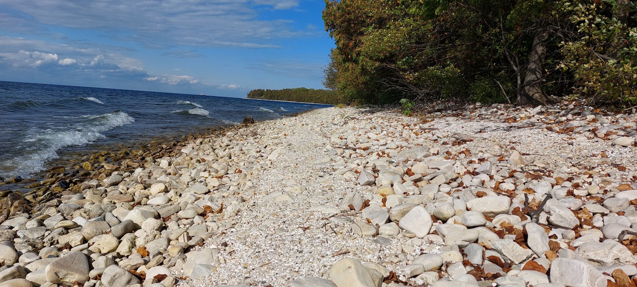 White rocky beach next to blue water with waves and green trees. - Eagle Bluff Lighthouse – Eagle Panorama loop from Greenwood Supper Club