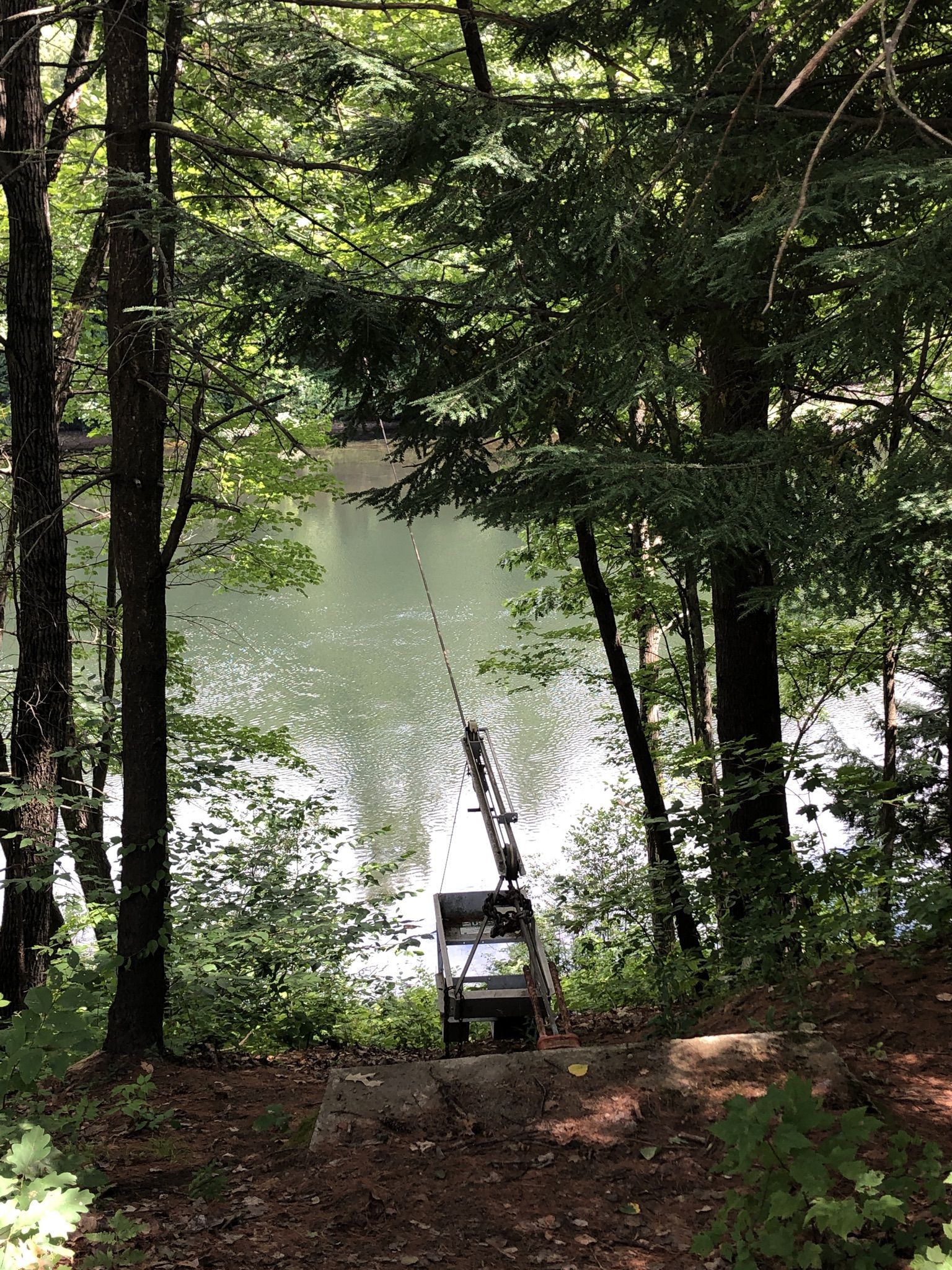 Fishing rod and cart on a shaded, wooded bank overlooking a river. - Road along Merrimack River loop from Boscawen
