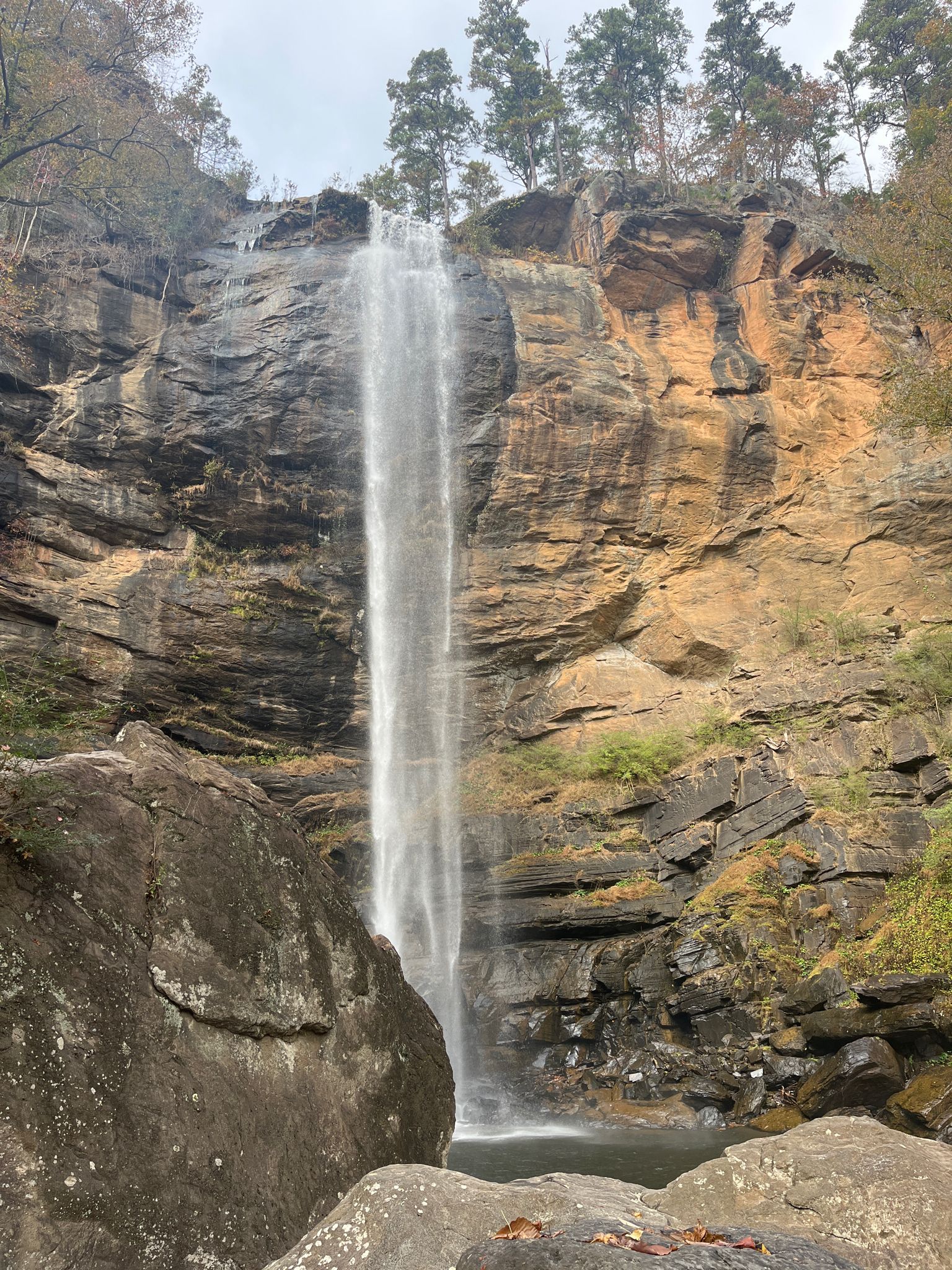 Tall waterfall cascades down a rocky cliff into a pool, surrounded by boulders. - Toccoa Falls