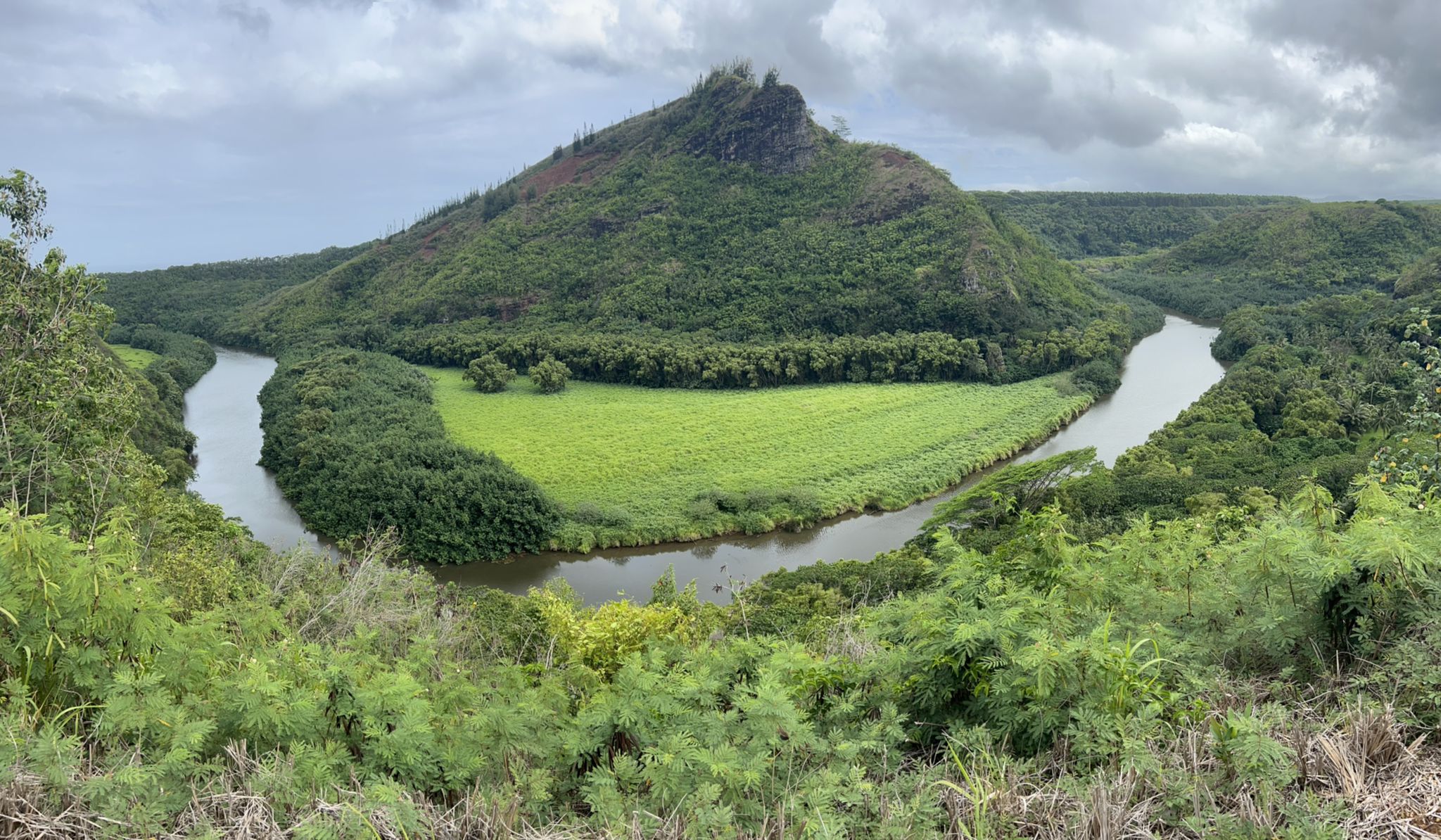 River winding around a lush green field and forested mountain under cloudy sky. - Ke Ala Hele Makalae Coastal Loop